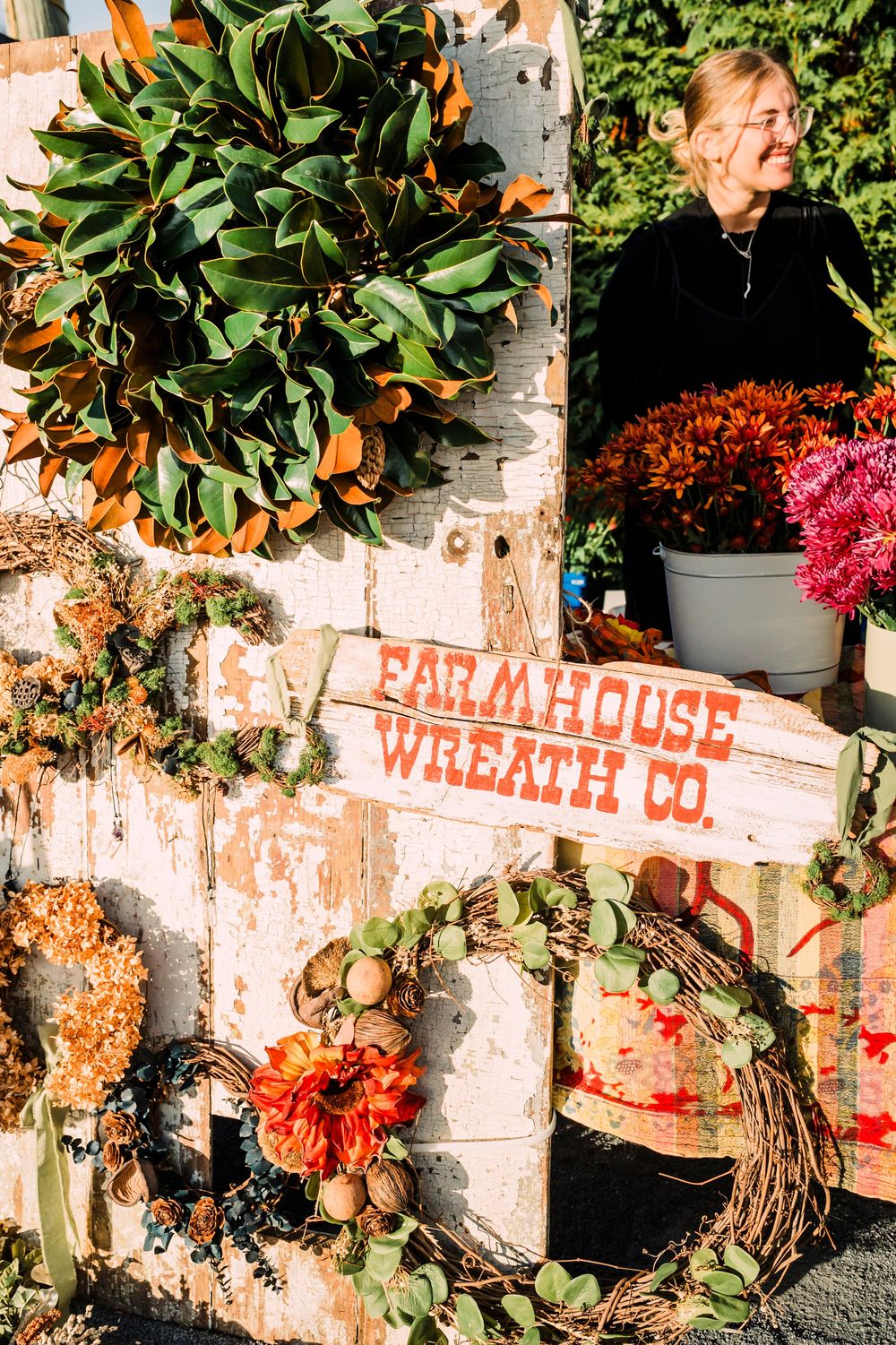 Rustic Farmhouse Wreath Co display with autumn wreaths, magnolia leaves and colorful floral arrangements in birch setting.