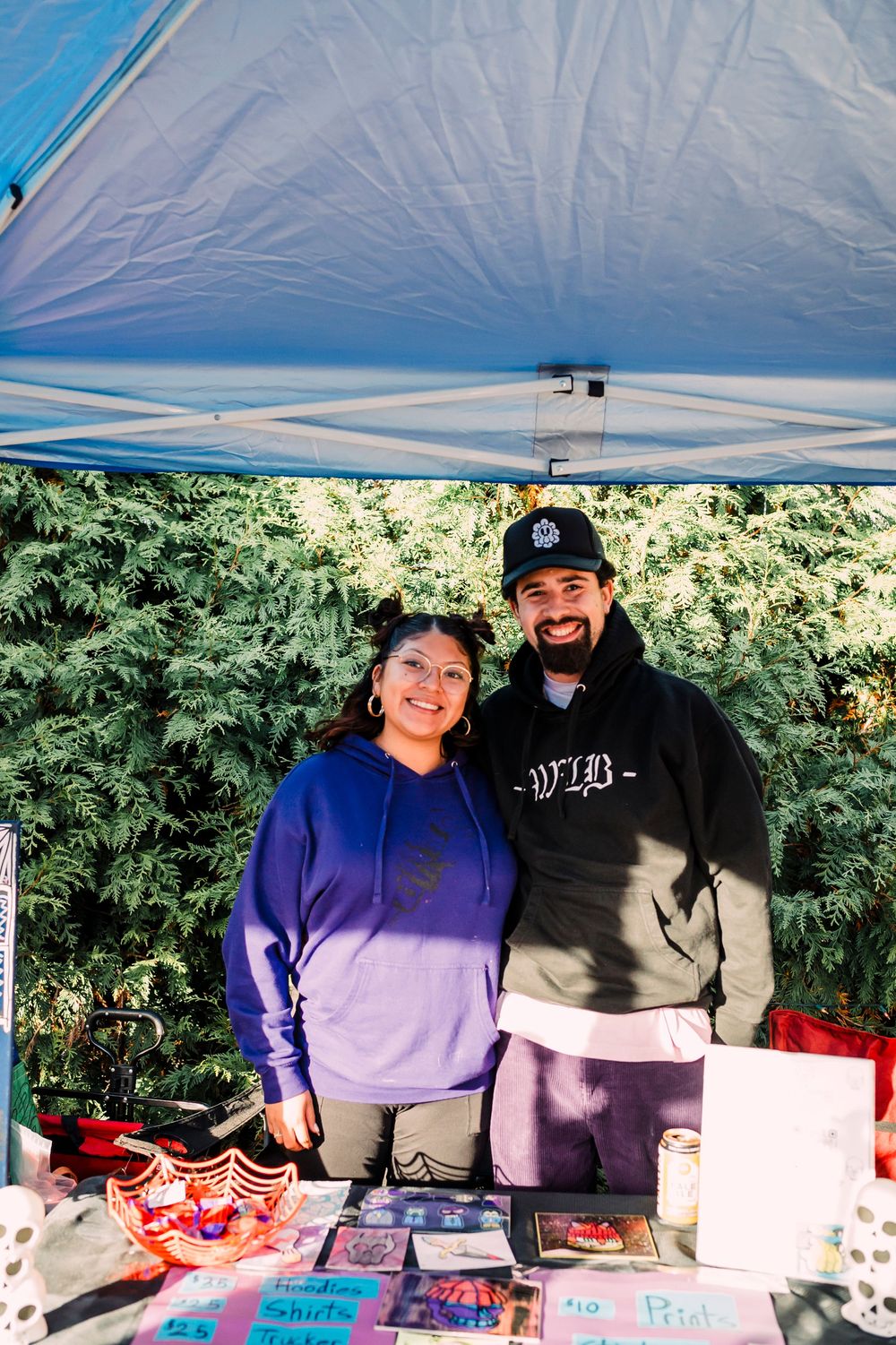 Two volunteers manage a merchandise booth under a blue canopy tent with various items displayed on a table.