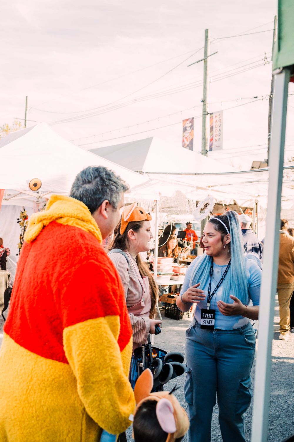 Group chats together at a street market wearing colorful clothing including a bright orange and yellow jacket.