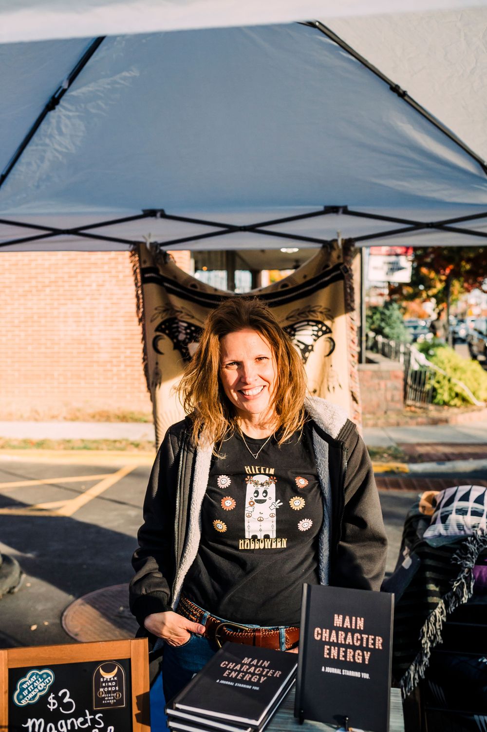 A vendor stands at their booth under a pop-up tent selling merchandise at an outdoor market.