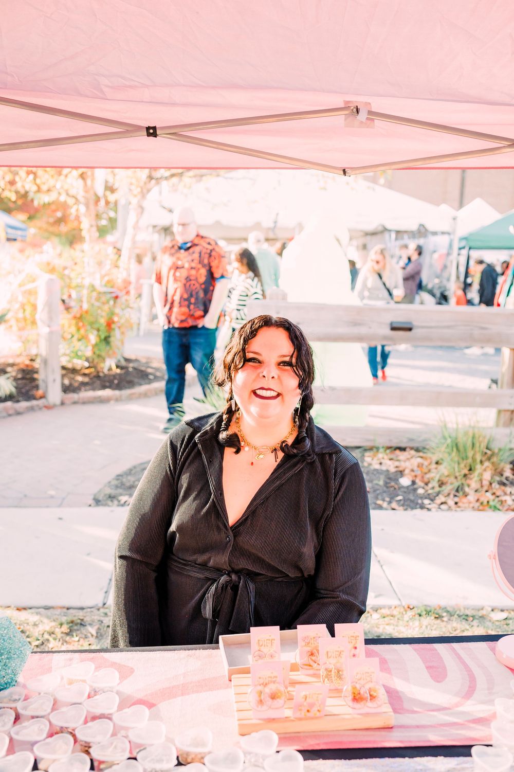 Vendor smiling behind booth display at outdoor market with pink signage and white tent overhead.