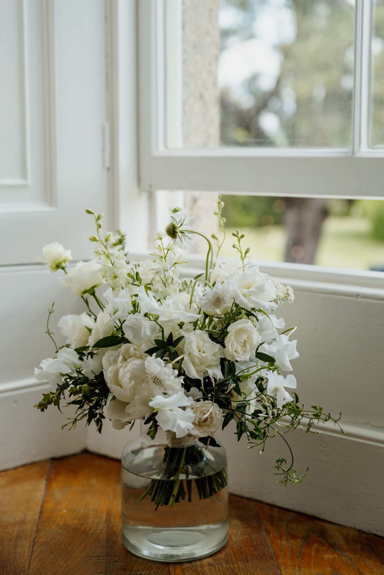 An elegant white rose and greenery flower arrangement in a glass vase sits on a wooden windowsill in natural light.