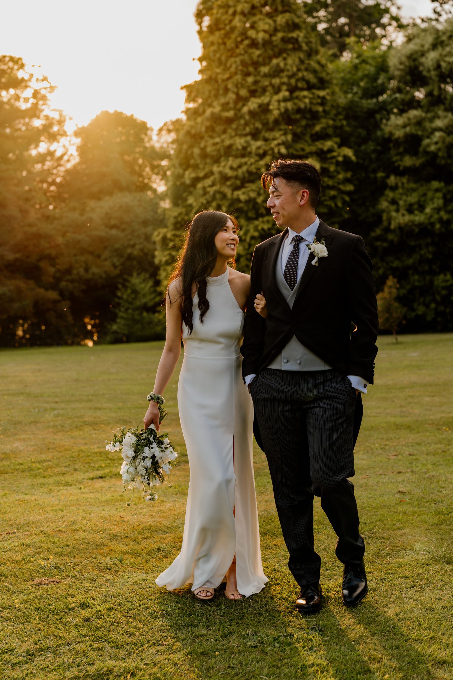 Wedding couple walks together at sunset on manicured lawn with golden light filtering through trees.