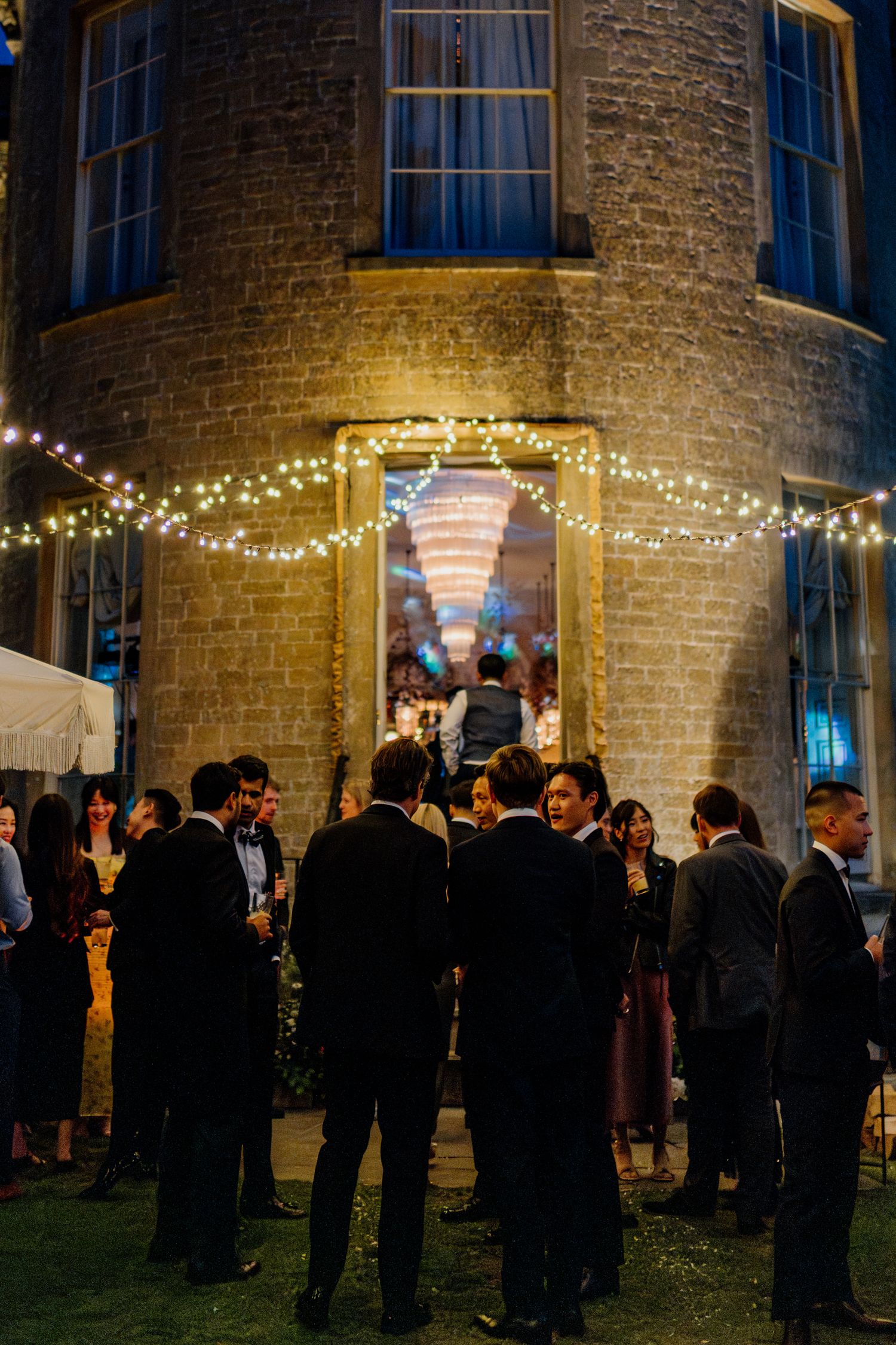 Evening wedding reception outside brick building with string lights and mingling guests.
