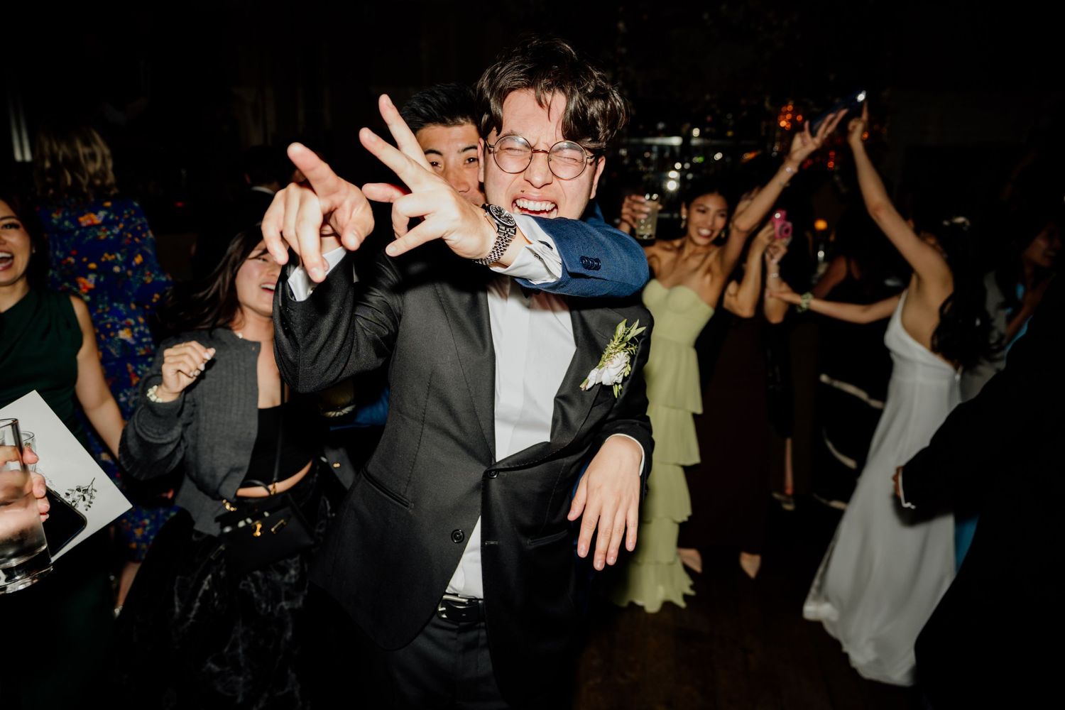 Wedding reception guests dance and celebrate in a dimly lit party atmosphere.