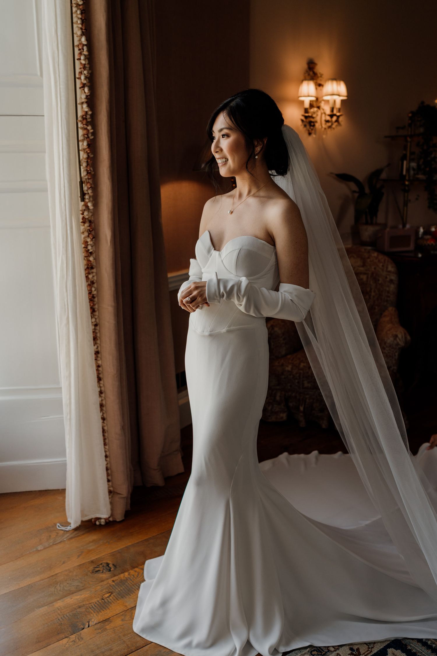 Elegant bride in fitted white gown and flowing veil poses in ornate room.