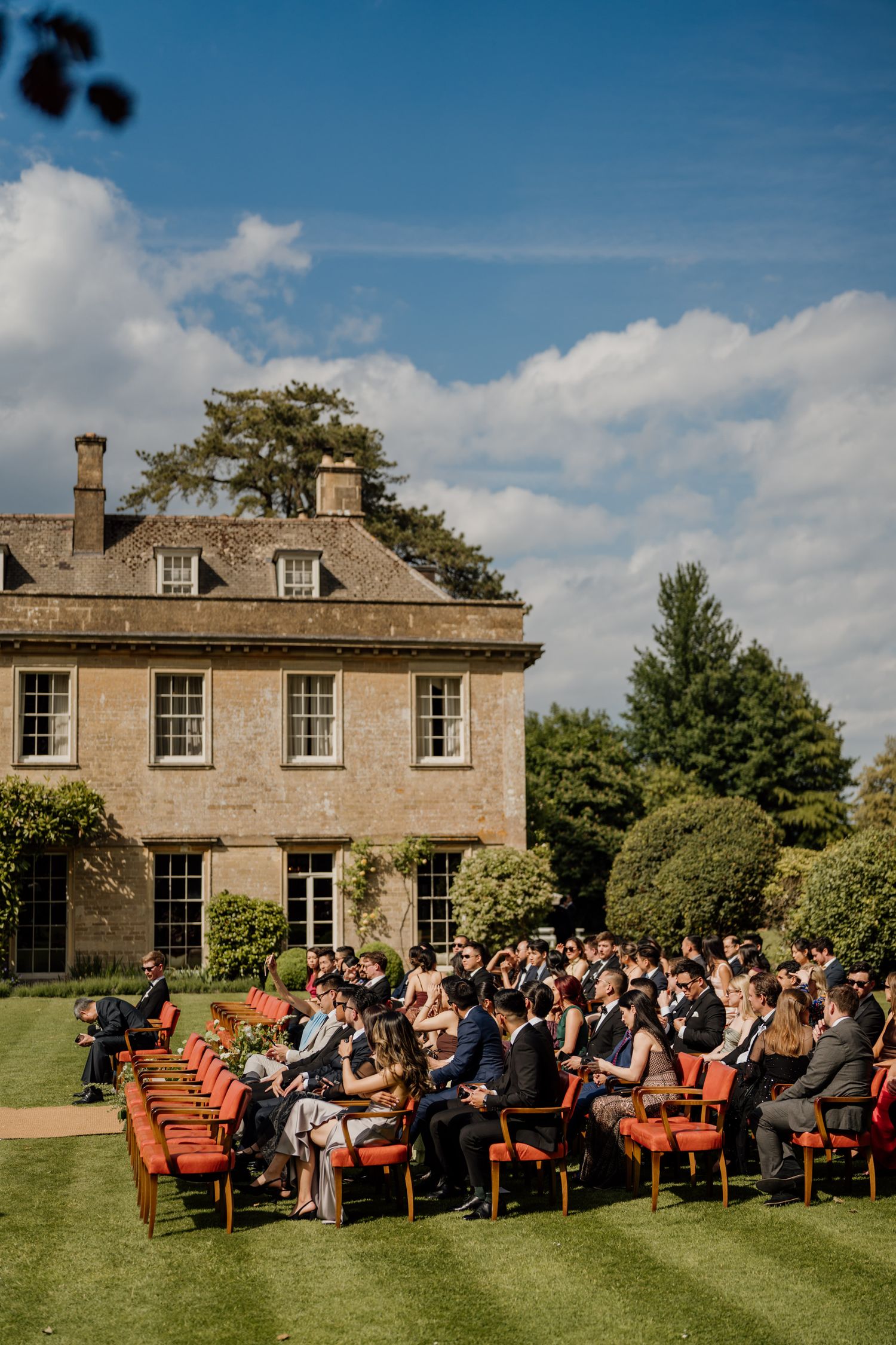Guests seated in red wooden chairs on a lawn for an outdoor ceremony in front of a historic stone mansion.