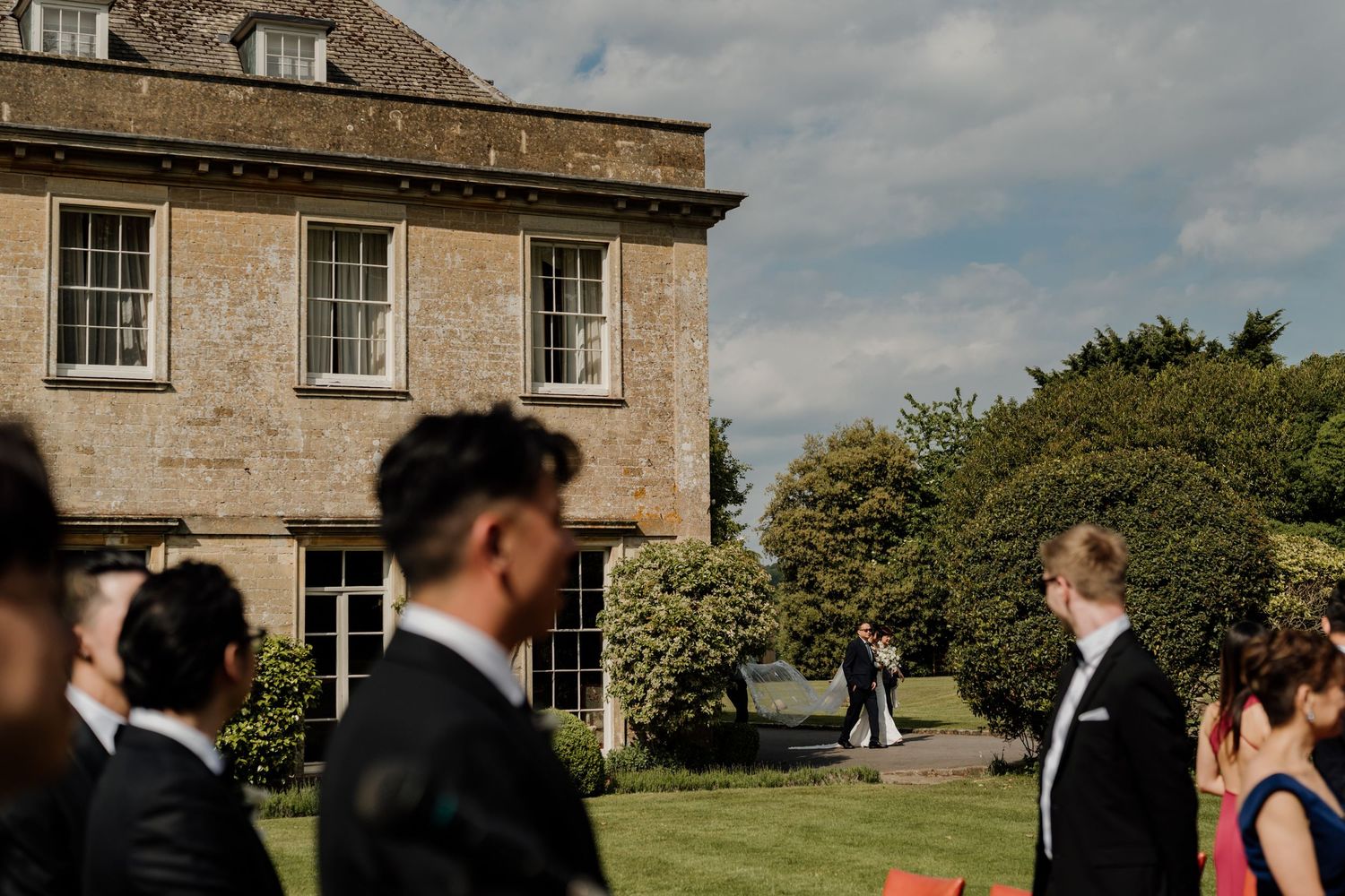 Wedding guests gathering outside a grand stone manor house on a sunny day.