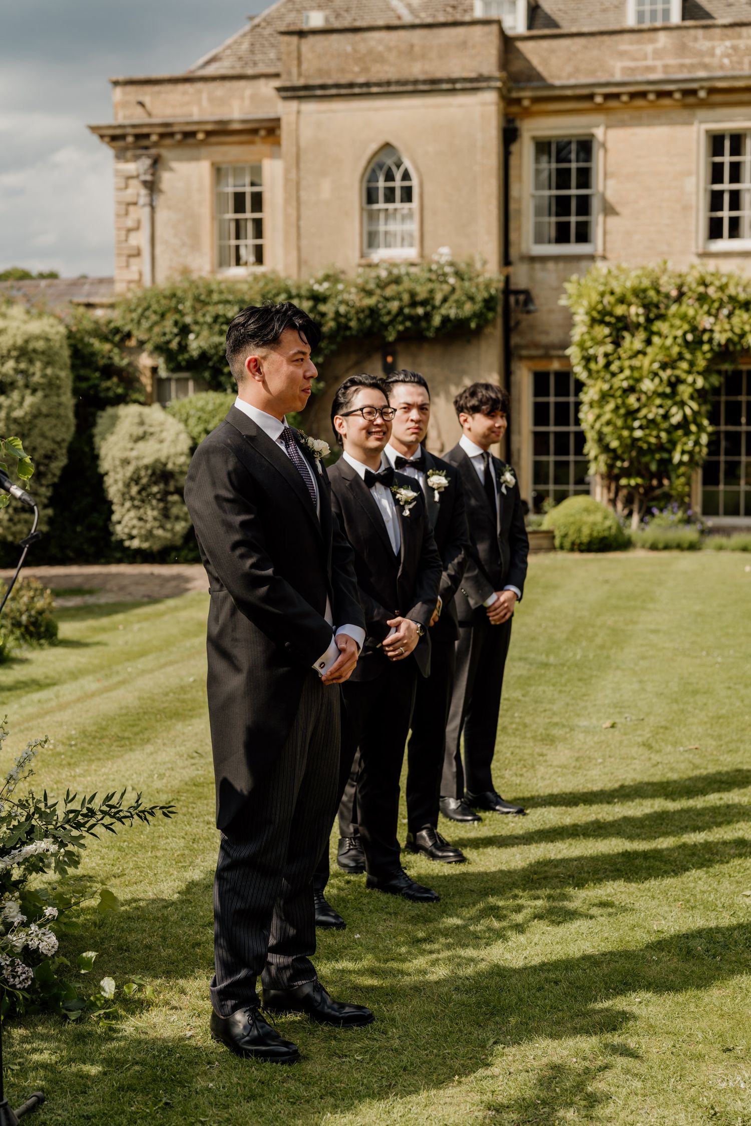A group of formally dressed attendants standing in a line on a manicured lawn outside an elegant stone building.