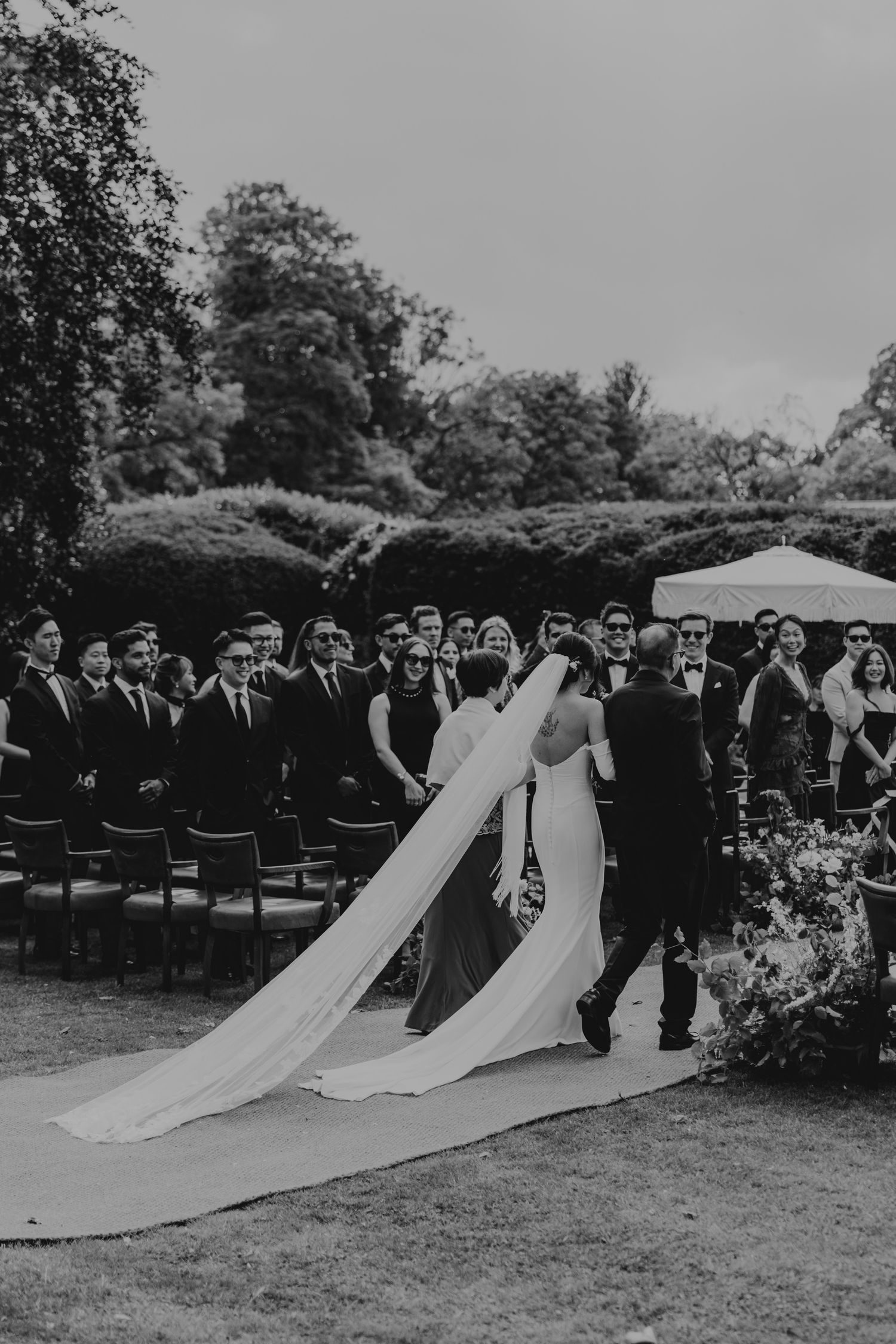 Large wedding party gatherers for a ceremony outdoors with long white gowns trailing on grass.