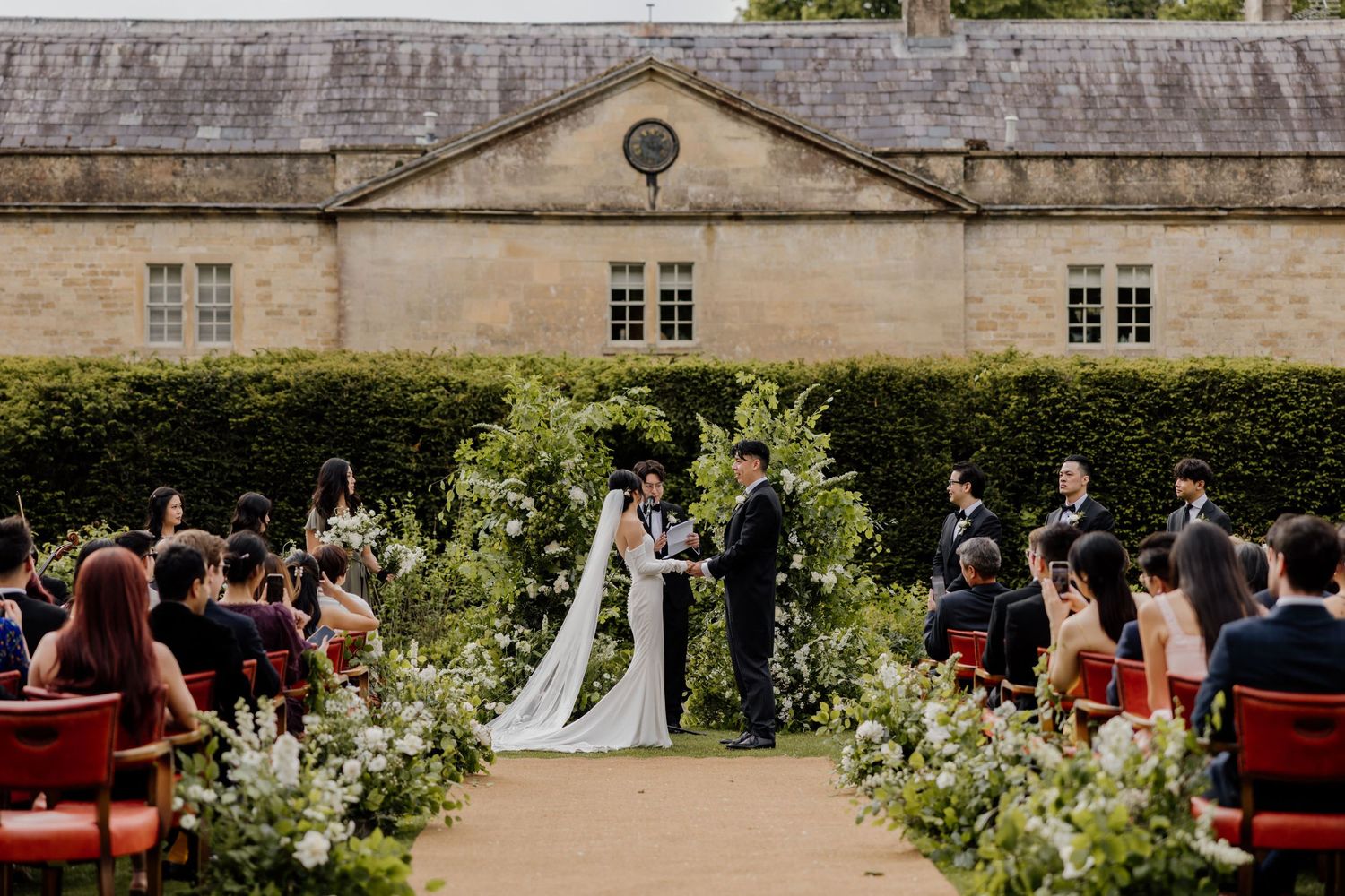 An outdoor garden wedding ceremony with red chairs and floral arrangements set against a stone building facade.