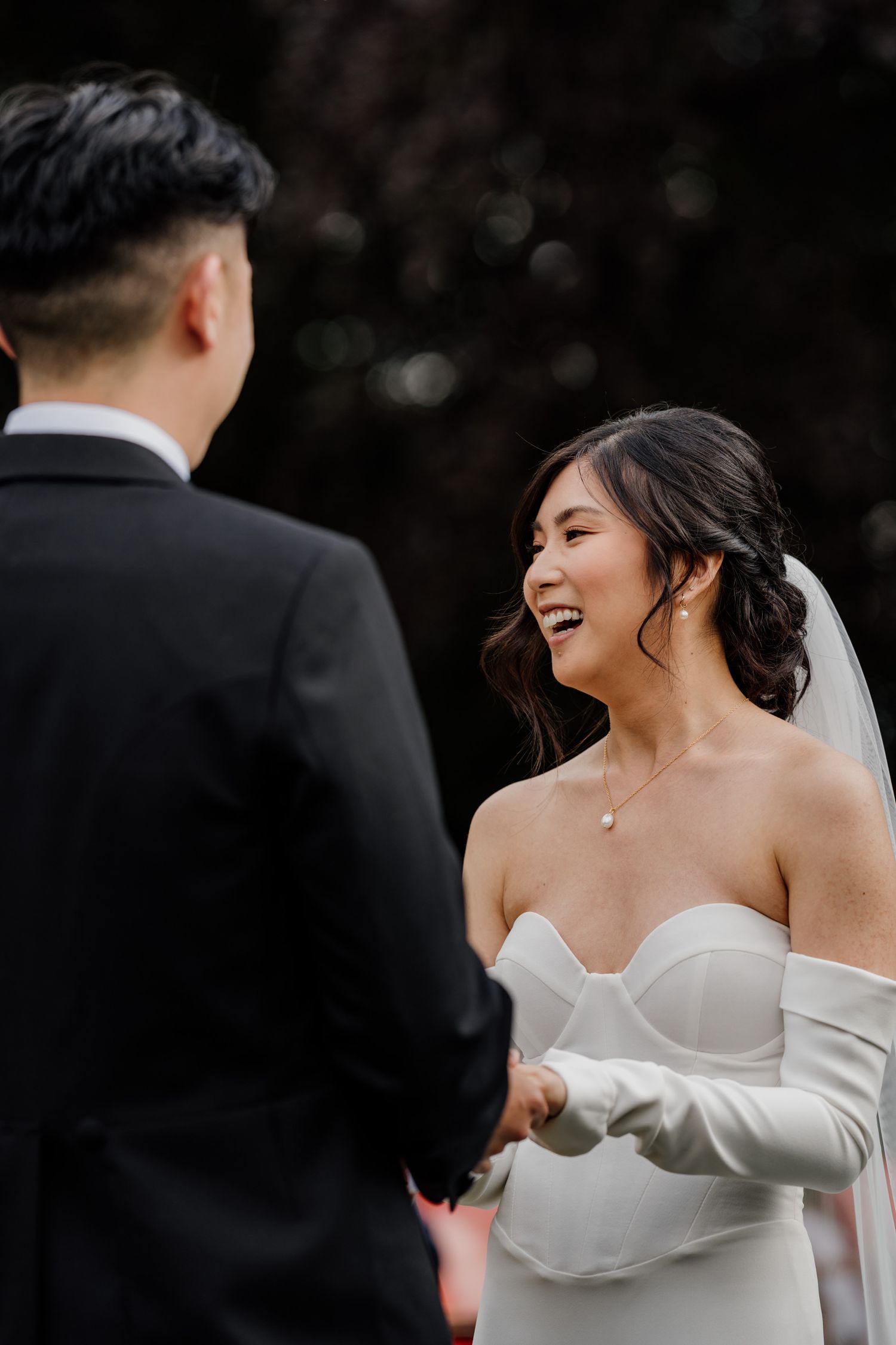 A bride laughs joyfully in her off-shoulder white wedding dress during an outdoor wedding ceremony.