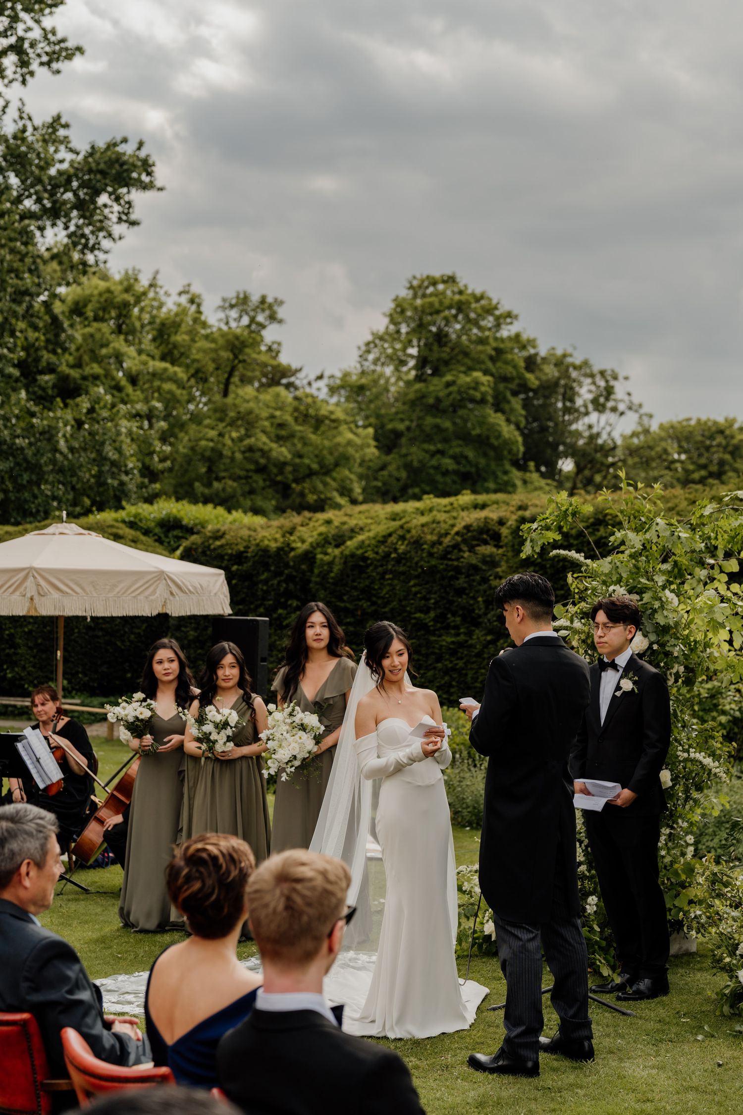 Outdoor wedding ceremony under cloudy skies with guests seated on a green lawn near a white tent.