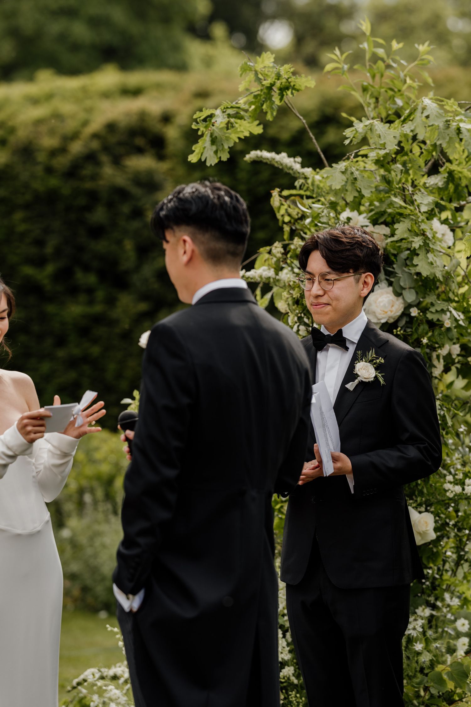 Wedding party members in formal attire standing among green foliage outdoors.