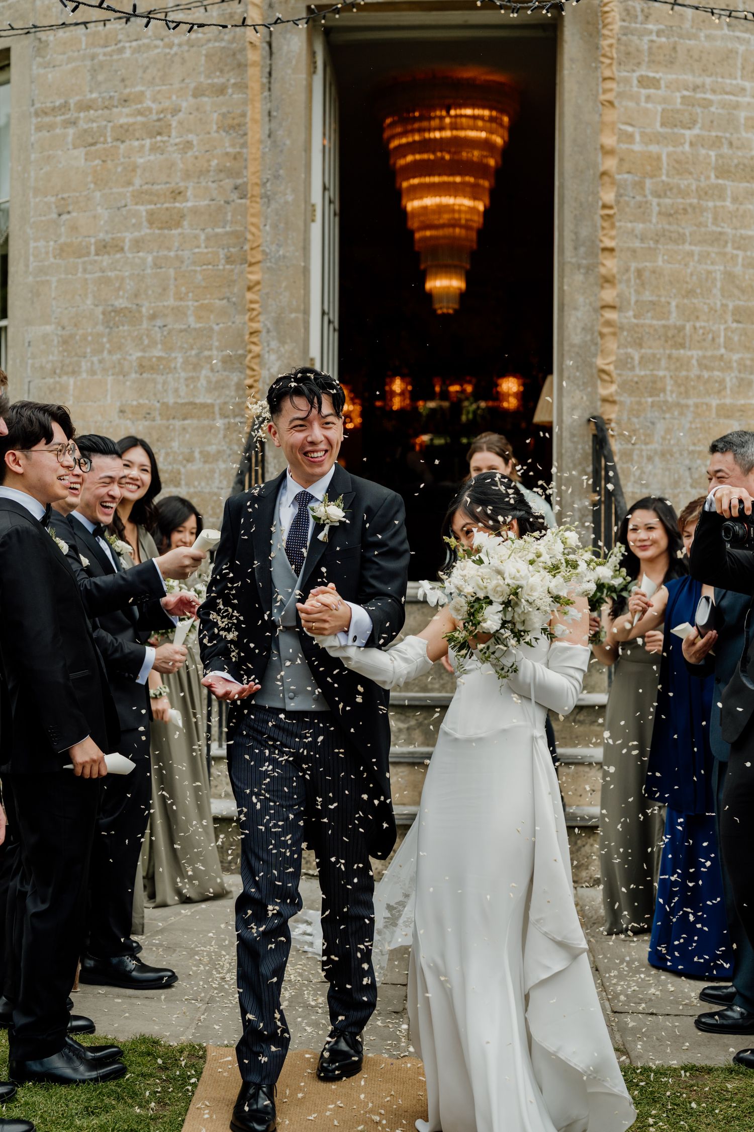 Newlyweds exit stone venue as guests shower them with confetti during daytime celebration.