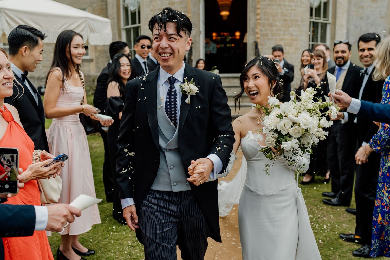 Wedding party and guests celebrating joyfully on the lawn outside a brick building.