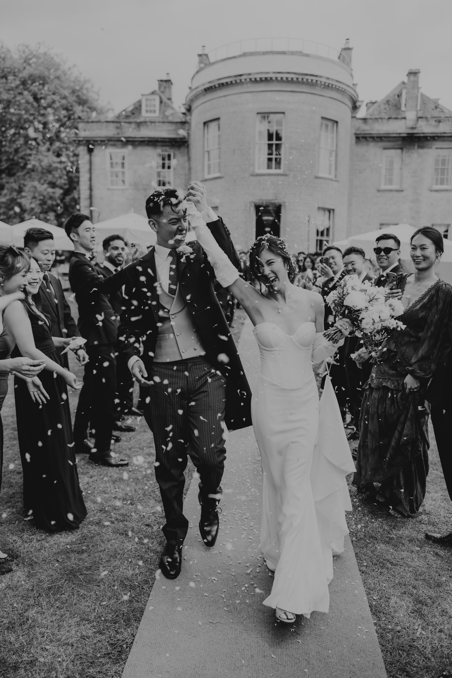 Black and white photo of wedding guests tossing confetti during an outdoor celebration at a historic venue.