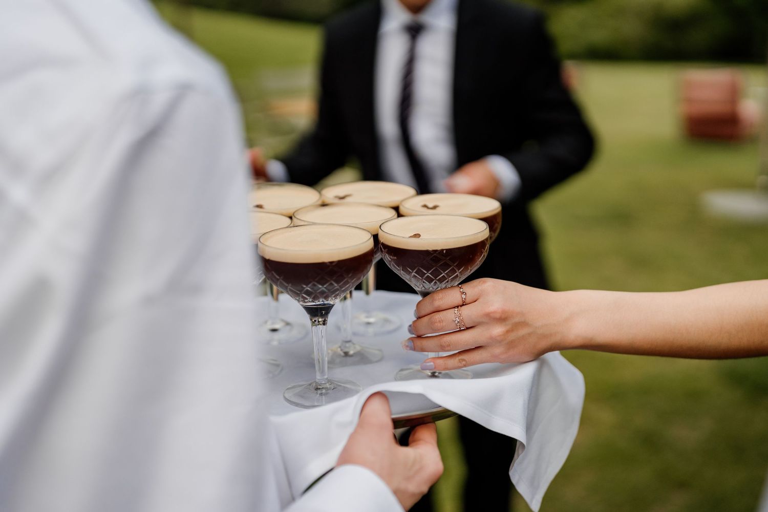 Server carries tray of espresso martinis on elegant glass stemware at outdoor garden wedding reception.
