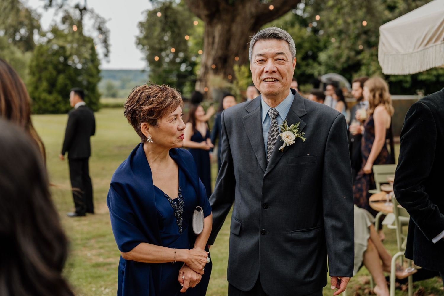 An outdoor wedding guest wearing a boutonniere stands next to someone in a navy shawl at an elegant garden reception.