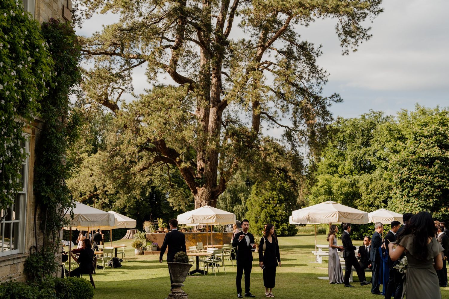 Wedding party gathers for photographs on the grounds of a historic estate surrounded by trees on a cloudy day.