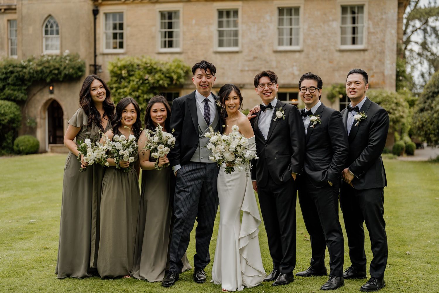 Wedding party poses together in formal attire on elegant manor house grounds with autumn trees in background.