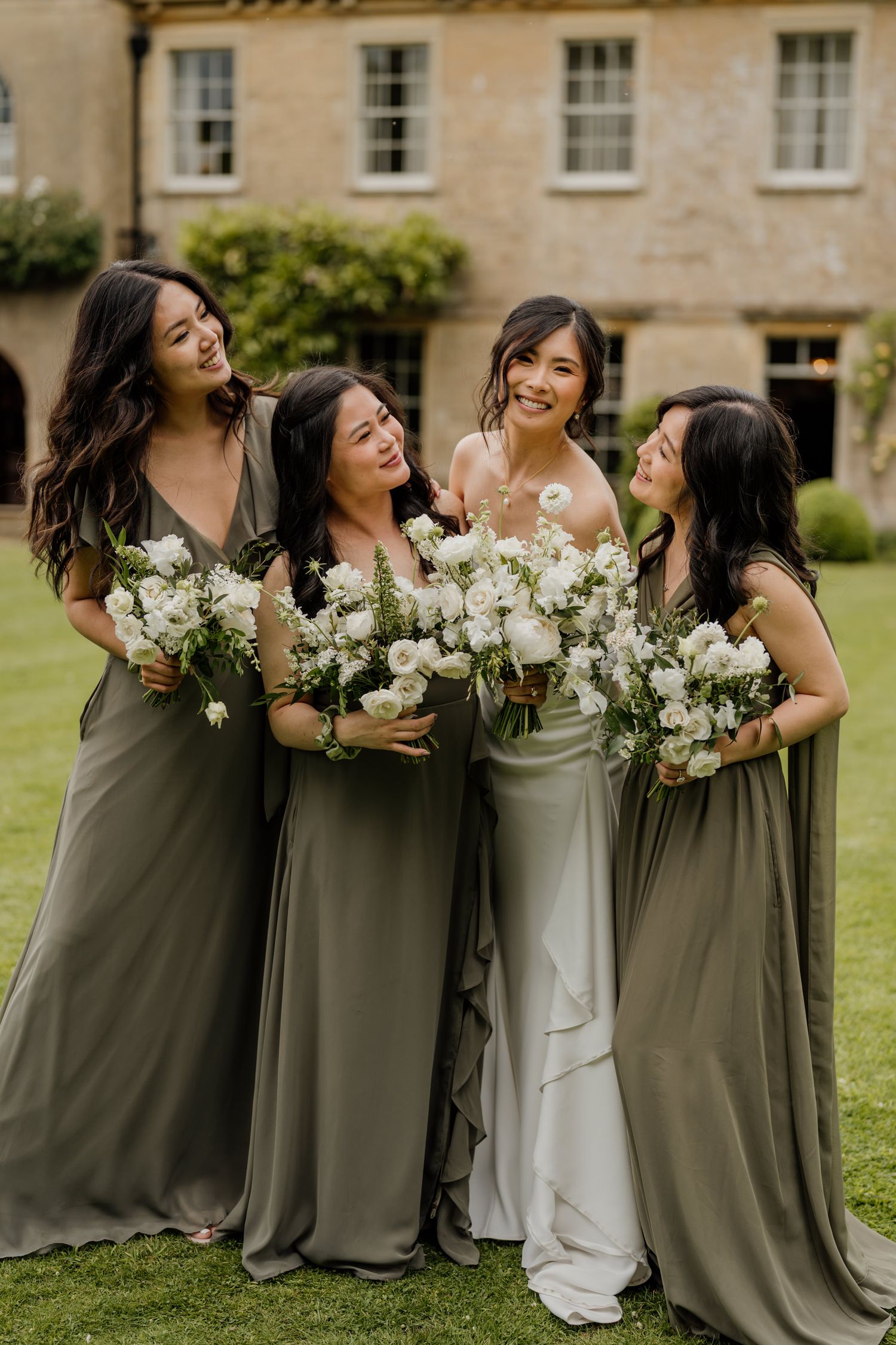 A bridal party in elegant sage green dresses laughs together while holding white bouquets on a grassy lawn.
