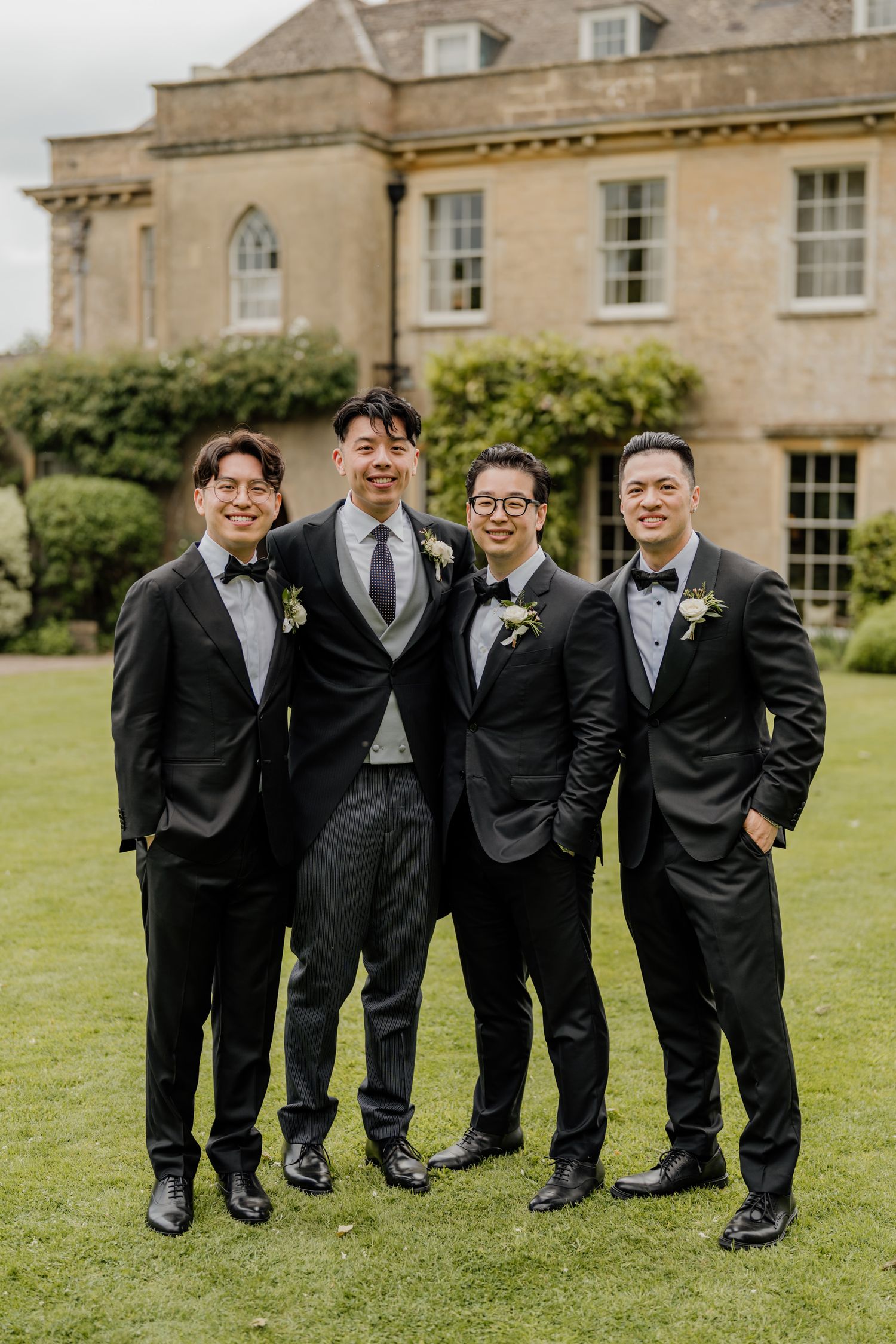 Groomsmen in black suits share laughs on a grand estate lawn.