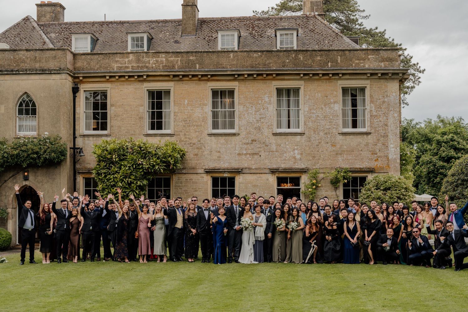 Large group photo of wedding guests gathered outside a grand stone manor house on overcast day.