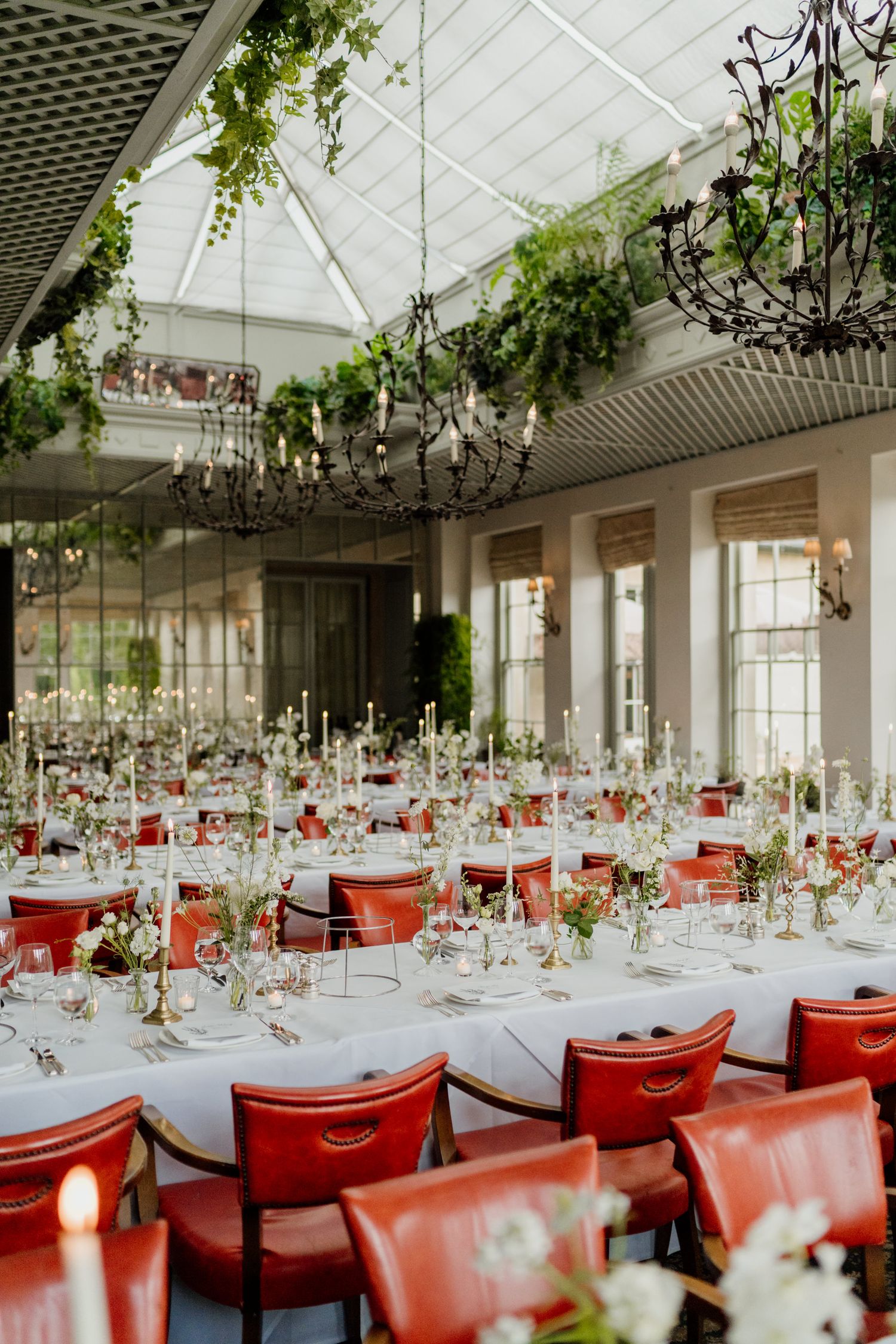 Elegant wedding reception venue with white linen tables, red chairs, and hanging greenery under a glass ceiling.