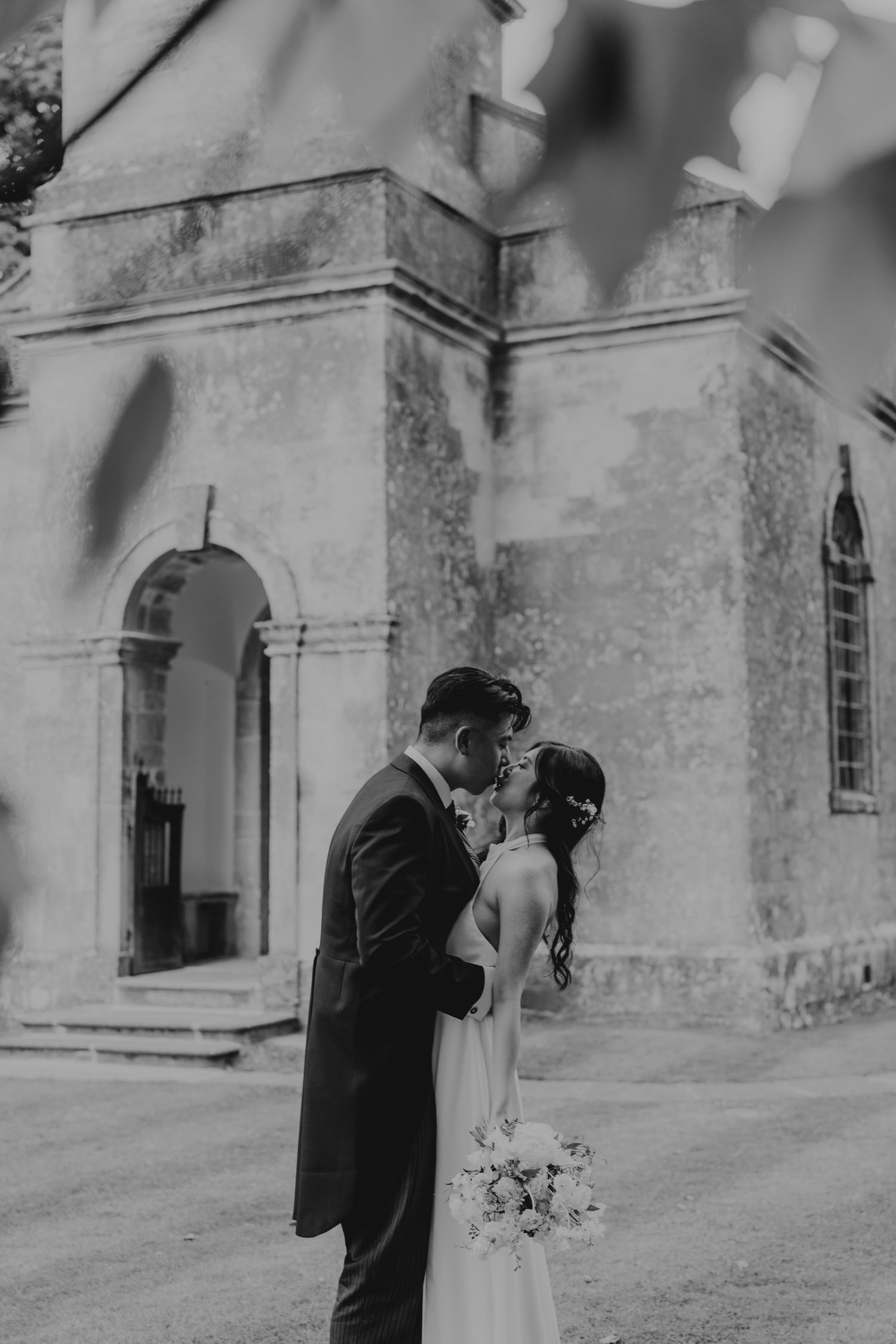 Black and white wedding portraits of a couple embracing on the grounds of a historic stone church.