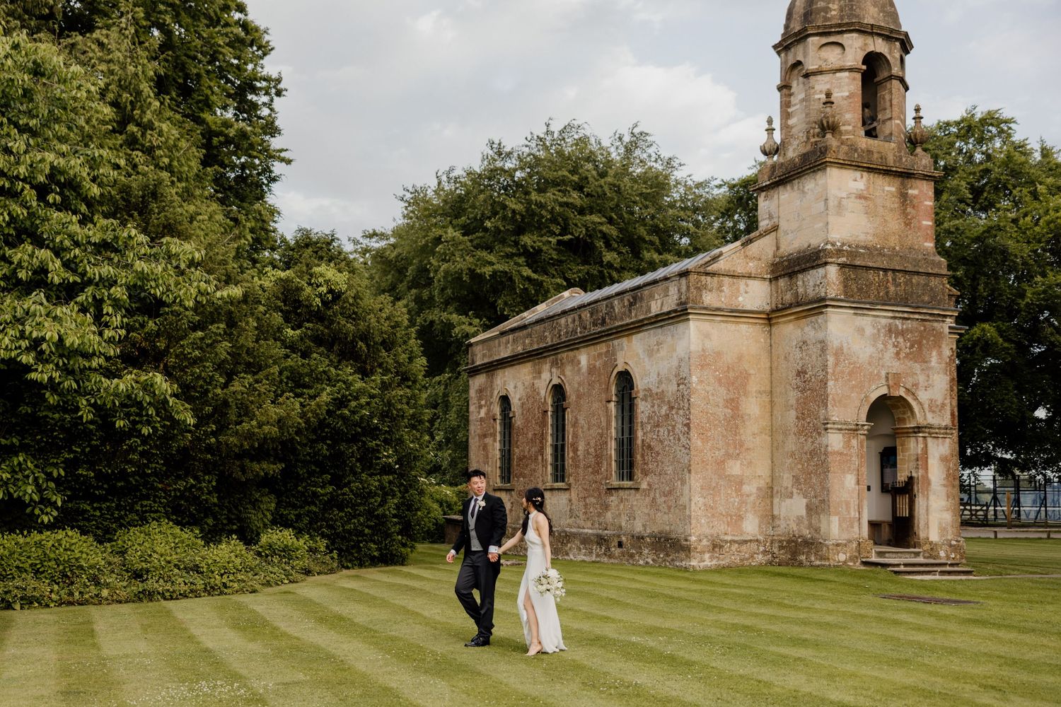 Newlyweds walk past a historic stone church on a grassy lawn during their wedding day celebrations.