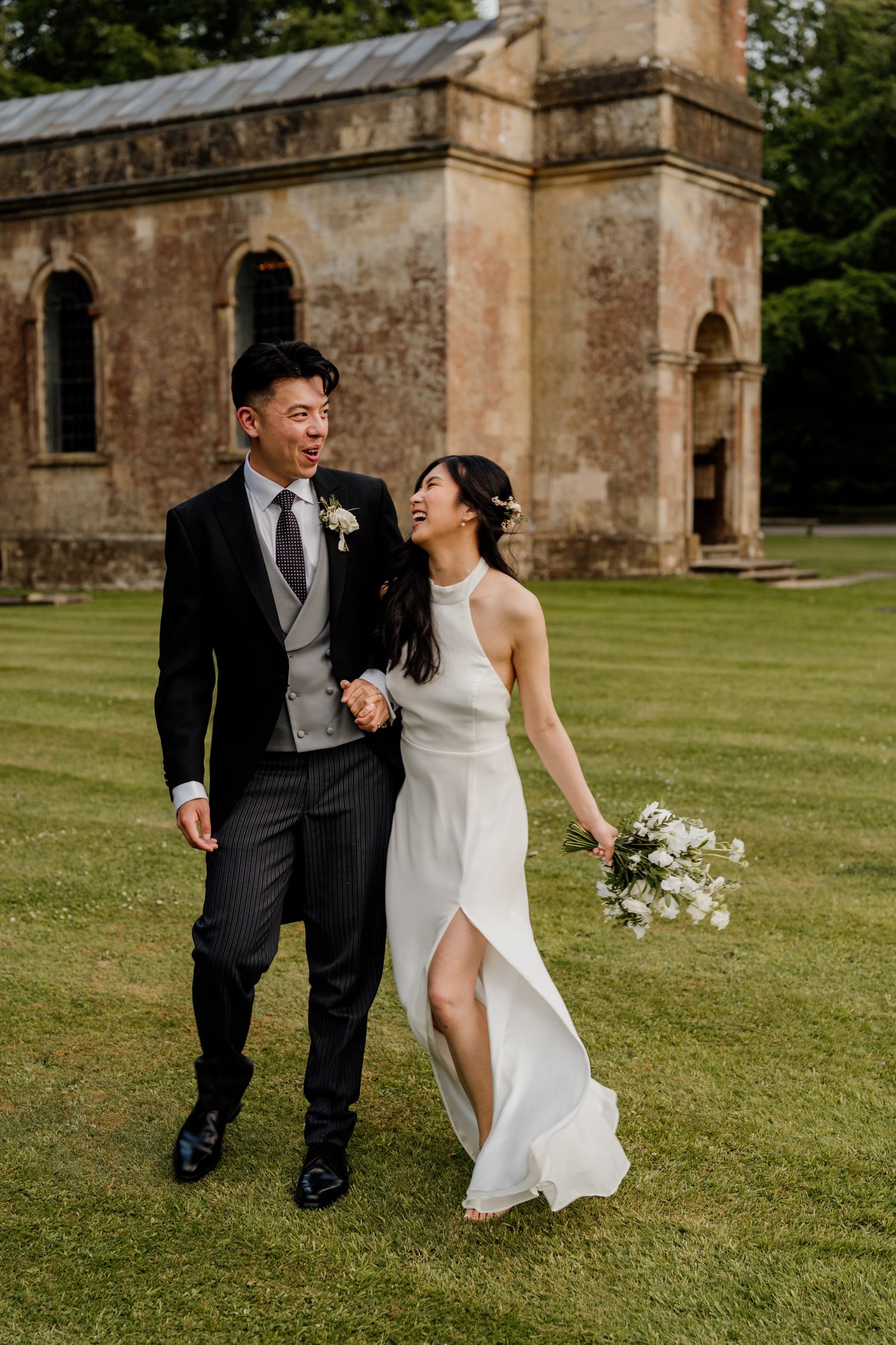 A couple shares romantic moments walking together on lush grounds near a stone building.