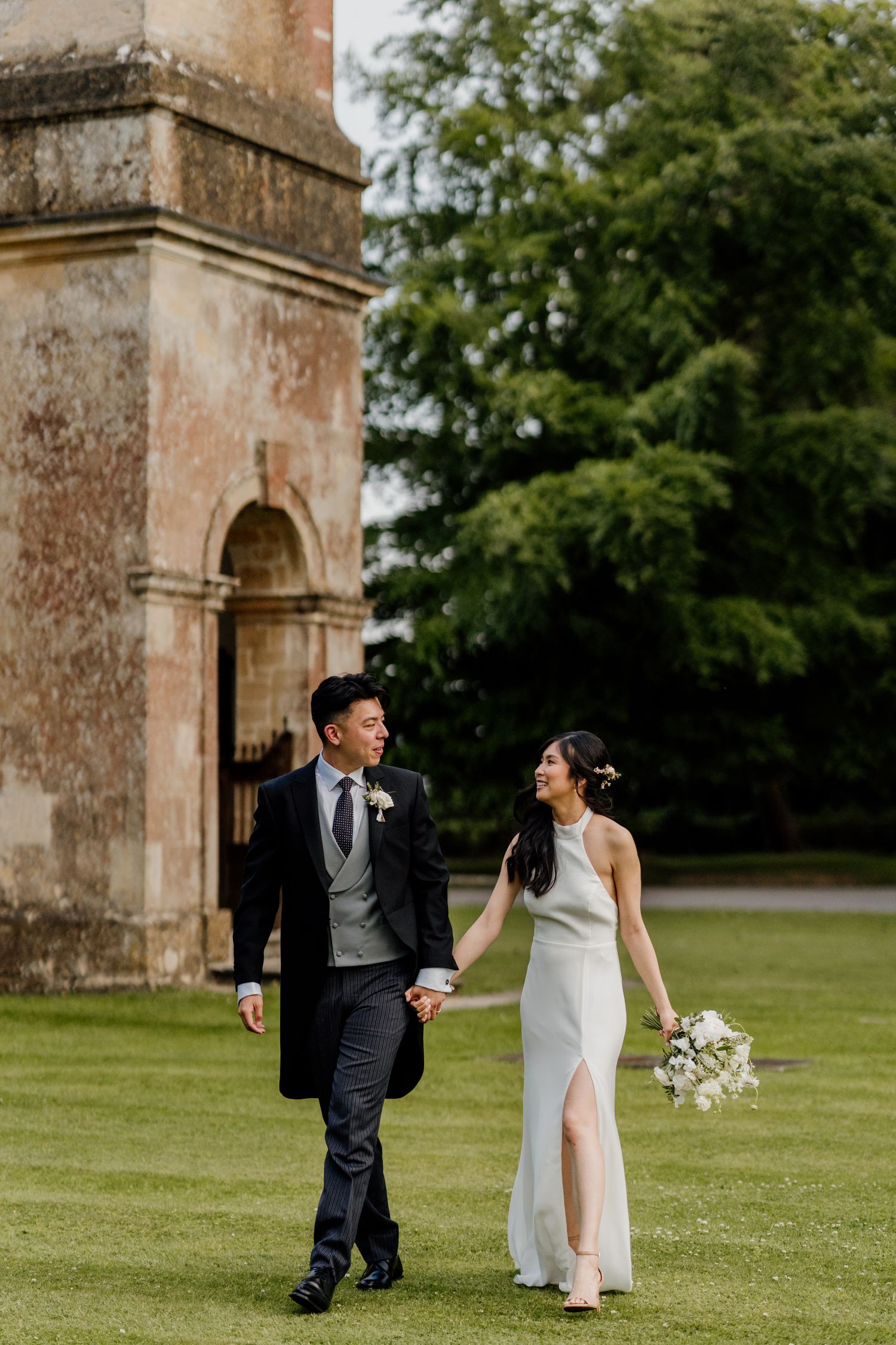 Elegant couple stroll hand in hand on manicured lawn in front of historic stone building.