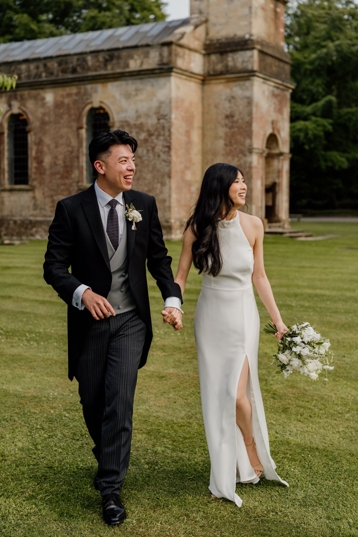 Newlyweds laugh and run playfully on the lawn of a historic stone church during their wedding photoshoot.