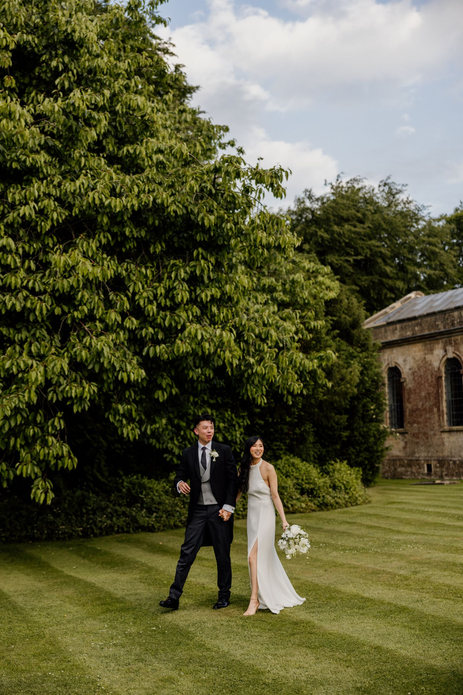 Wedding couple poses playfully on expansive lawn with historic building and lush trees in background.