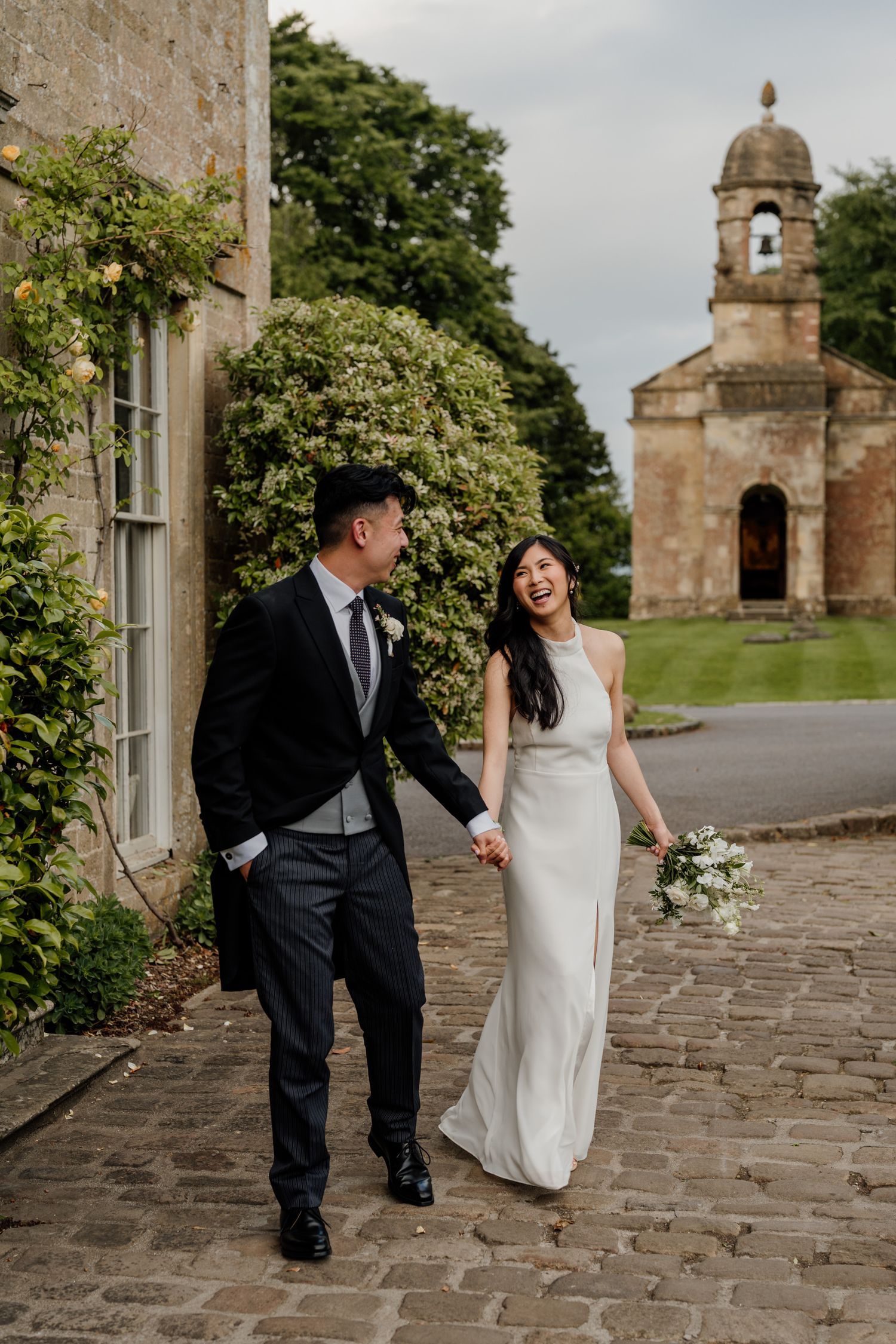 Wedding couple walking along a stone path with historic church building in the background.