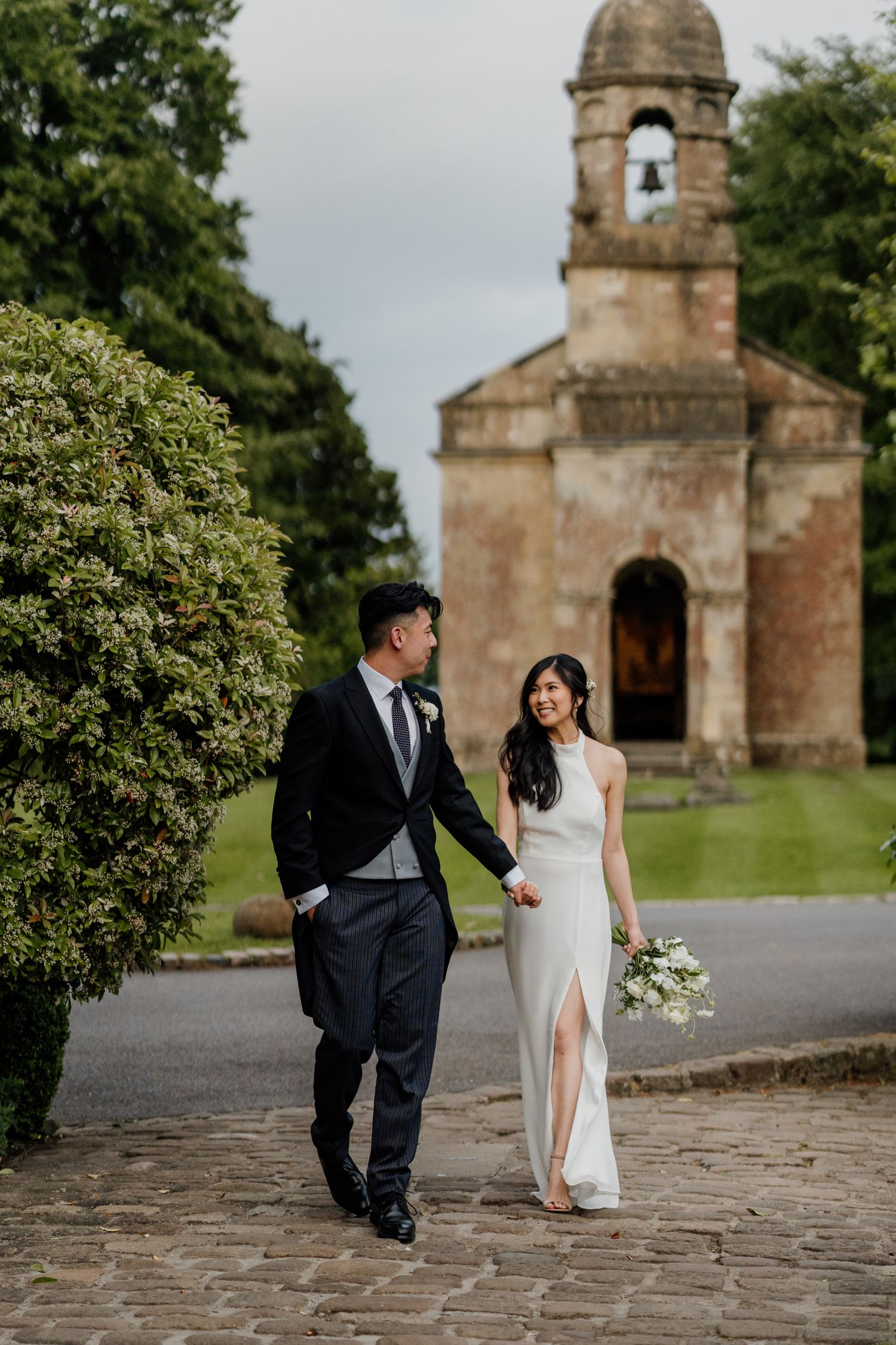 Wedding couple walks along stone path by historic church building.