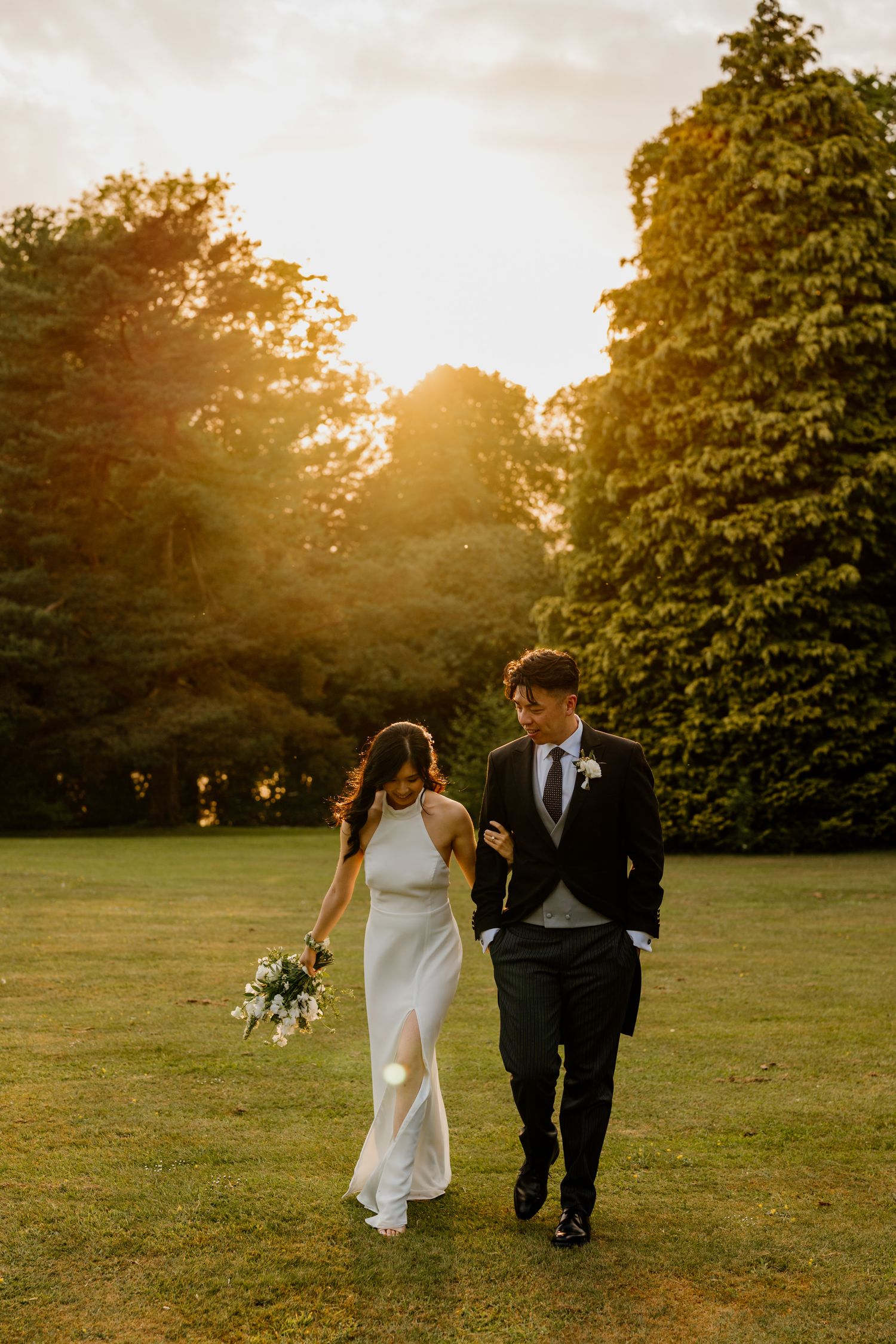 A couple walks hand in hand across a sunlit lawn during golden hour at their wedding.