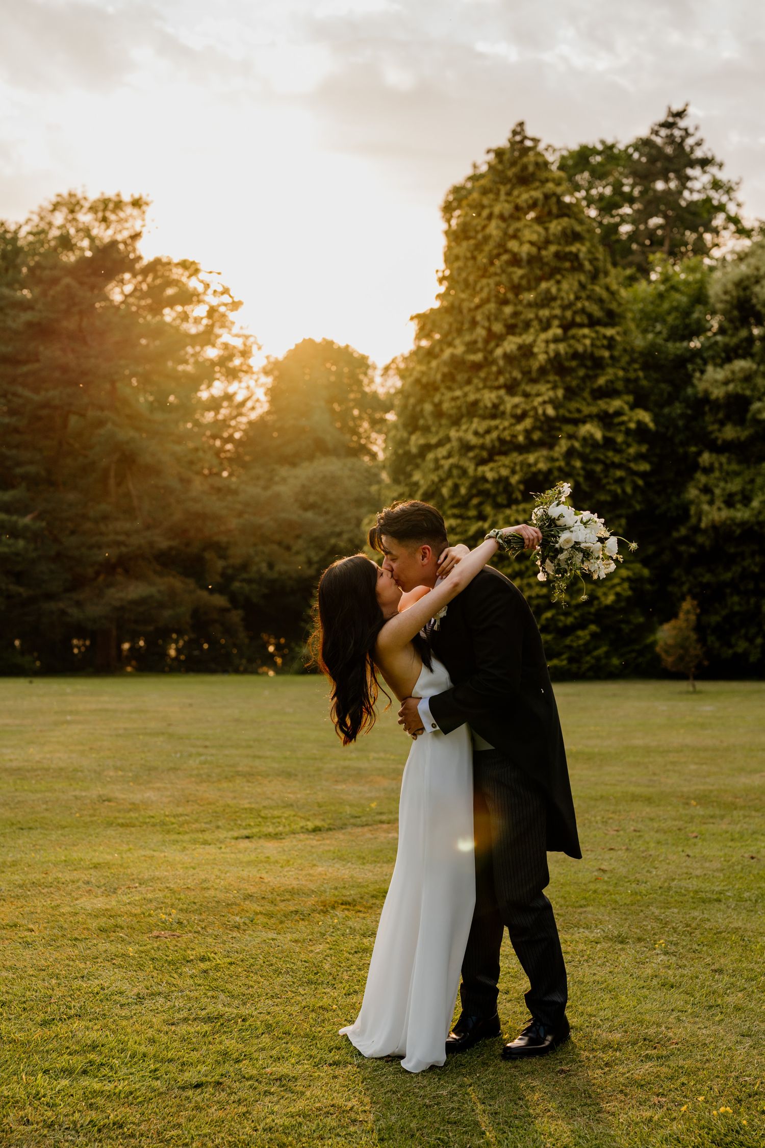 A couple in wedding attire dances together in a sunlit field during golden hour at sunset.