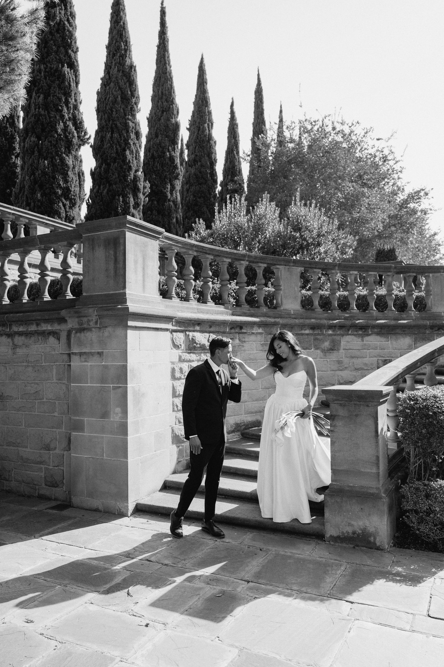 Black and white wedding series showing couple walking through a scenic garden terrace with cypress trees and stone balustrade.