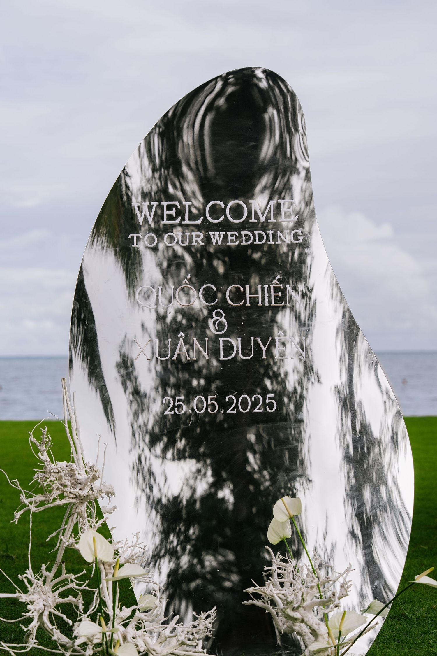 Reflective oval wedding welcome sign reading Quoc Chien and Xuan Duyen 25.05.2025 by the ocean.
