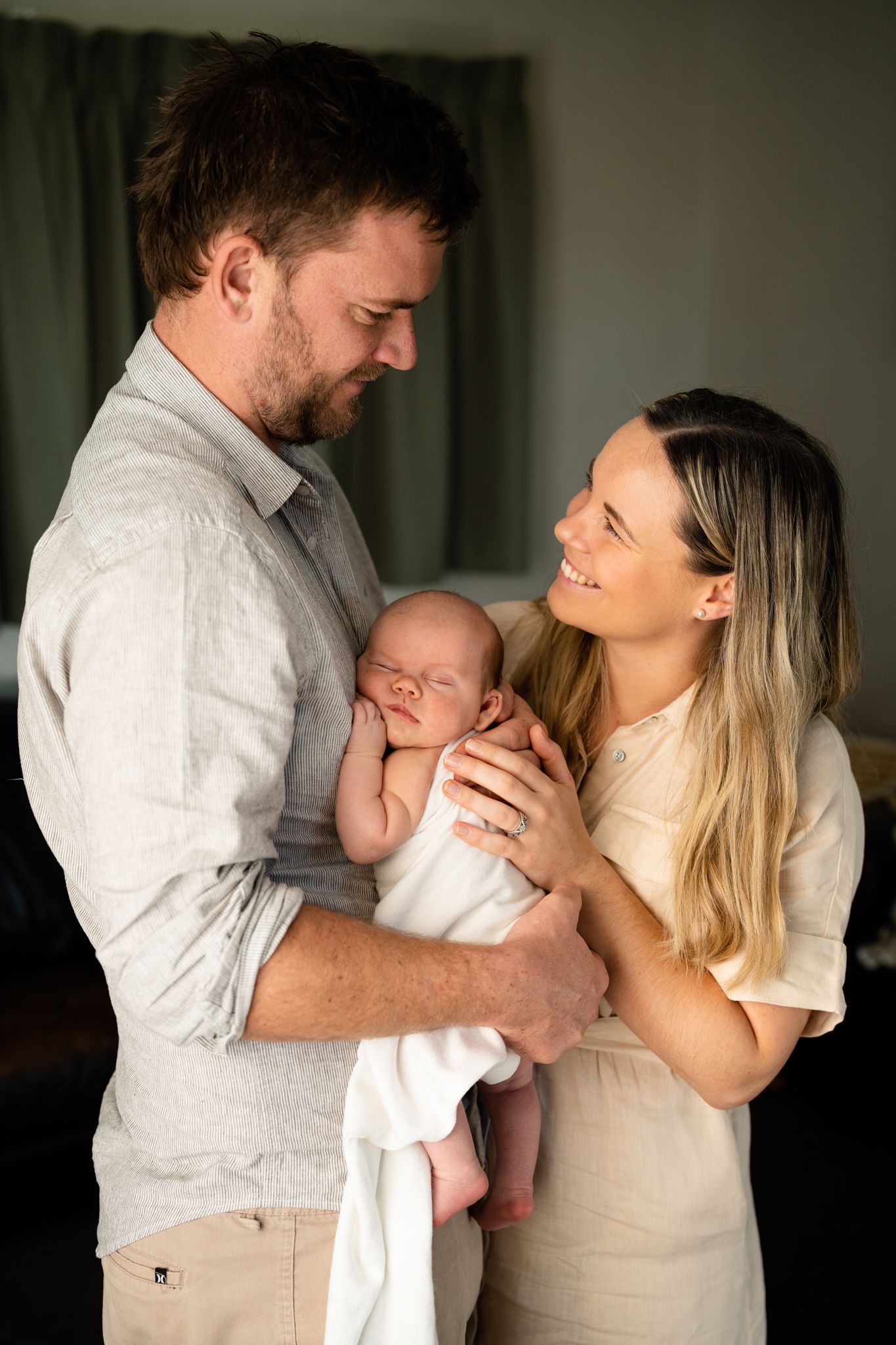 A couple in white clothing embraces tenderly while holding a newborn baby in a series of intimate indoor portraits.
