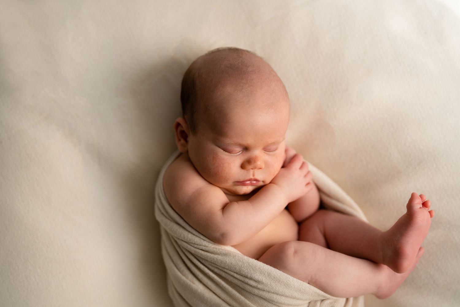 A peaceful sleeping newborn baby swaddled in soft white fabric rests on a cream colored background.
