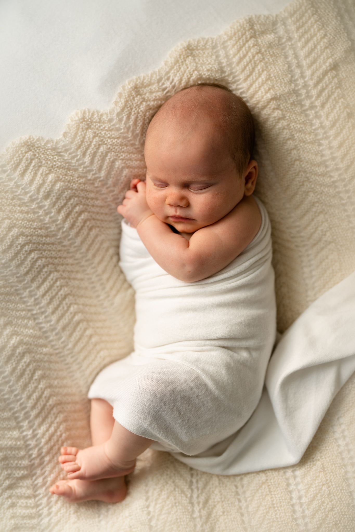 Sleeping baby wrapped in soft white swaddle blanket lying on cream chevron textured blanket.