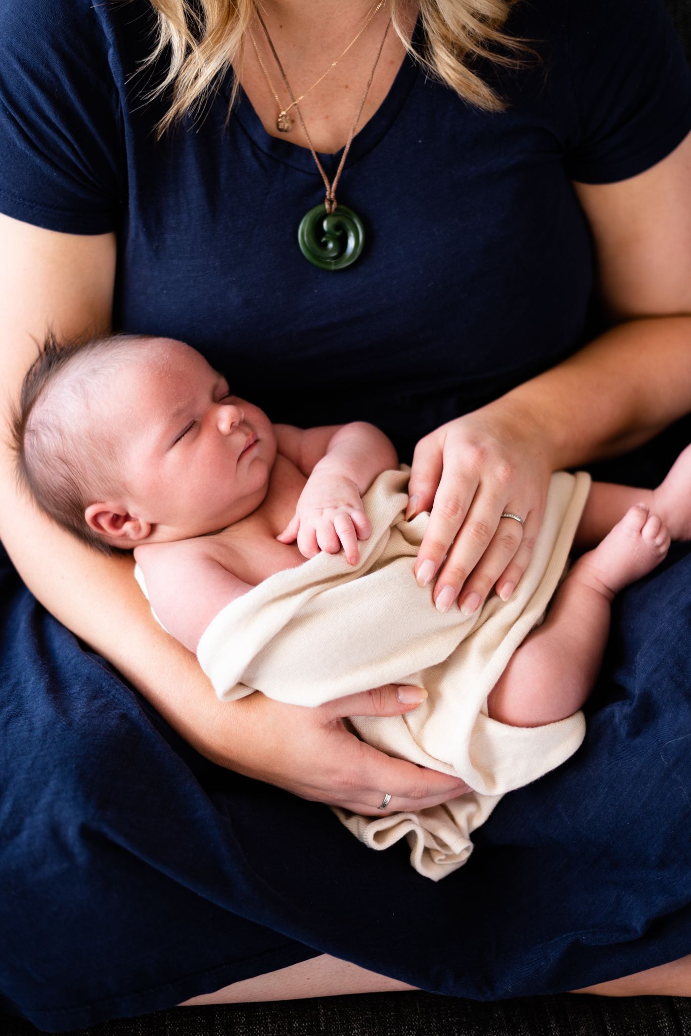 Newborn baby in cream wrap resting on parent wearing navy blue shirt with green pendant.