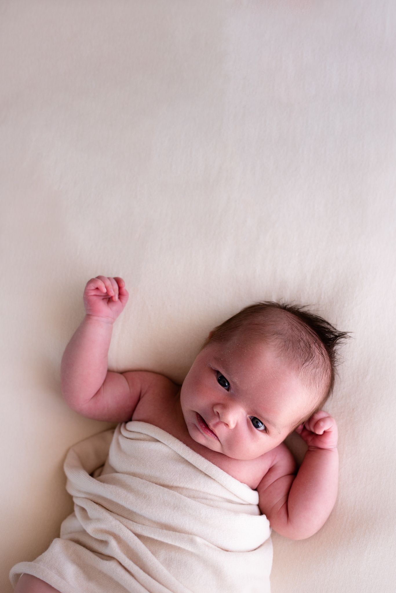 Newborn baby wrapped in white cloth lying on light background with arms raised.