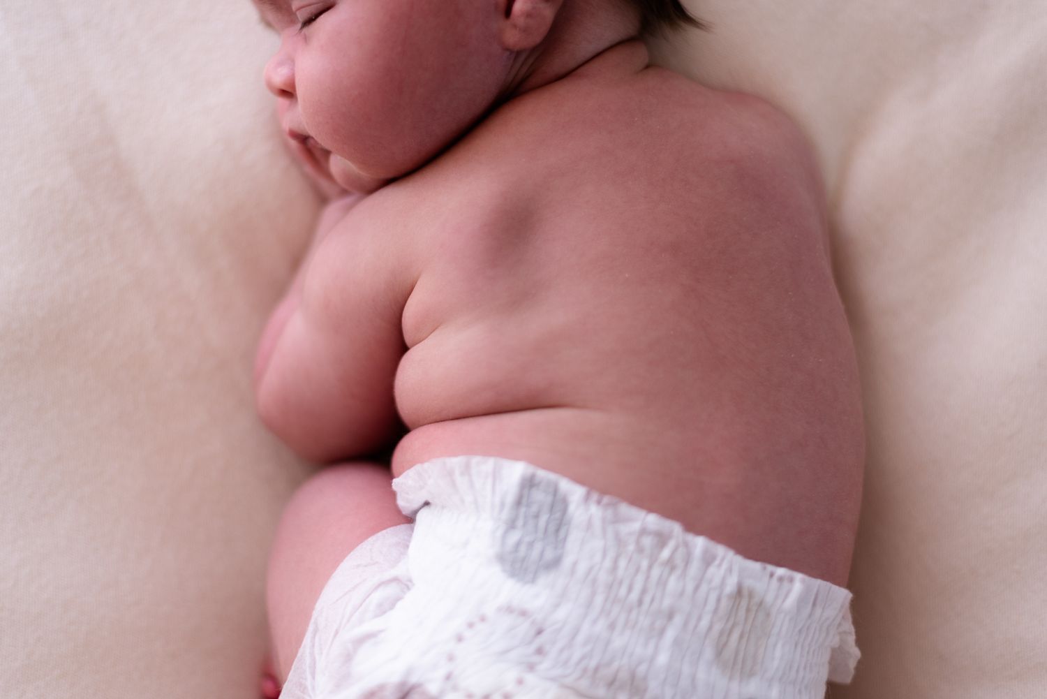 Sleeping baby in diaper resting on soft cream colored surface.