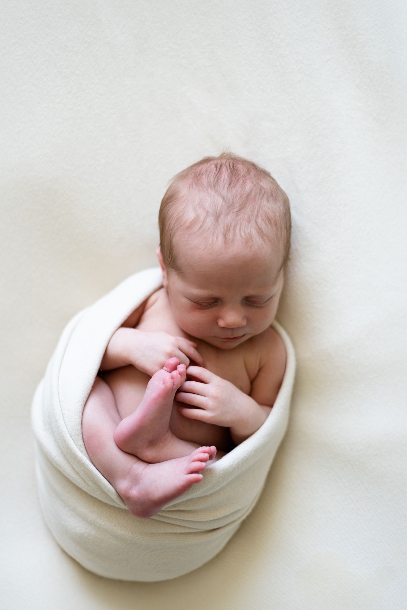 Newborn baby curled up in white wrap looking at tiny toes.