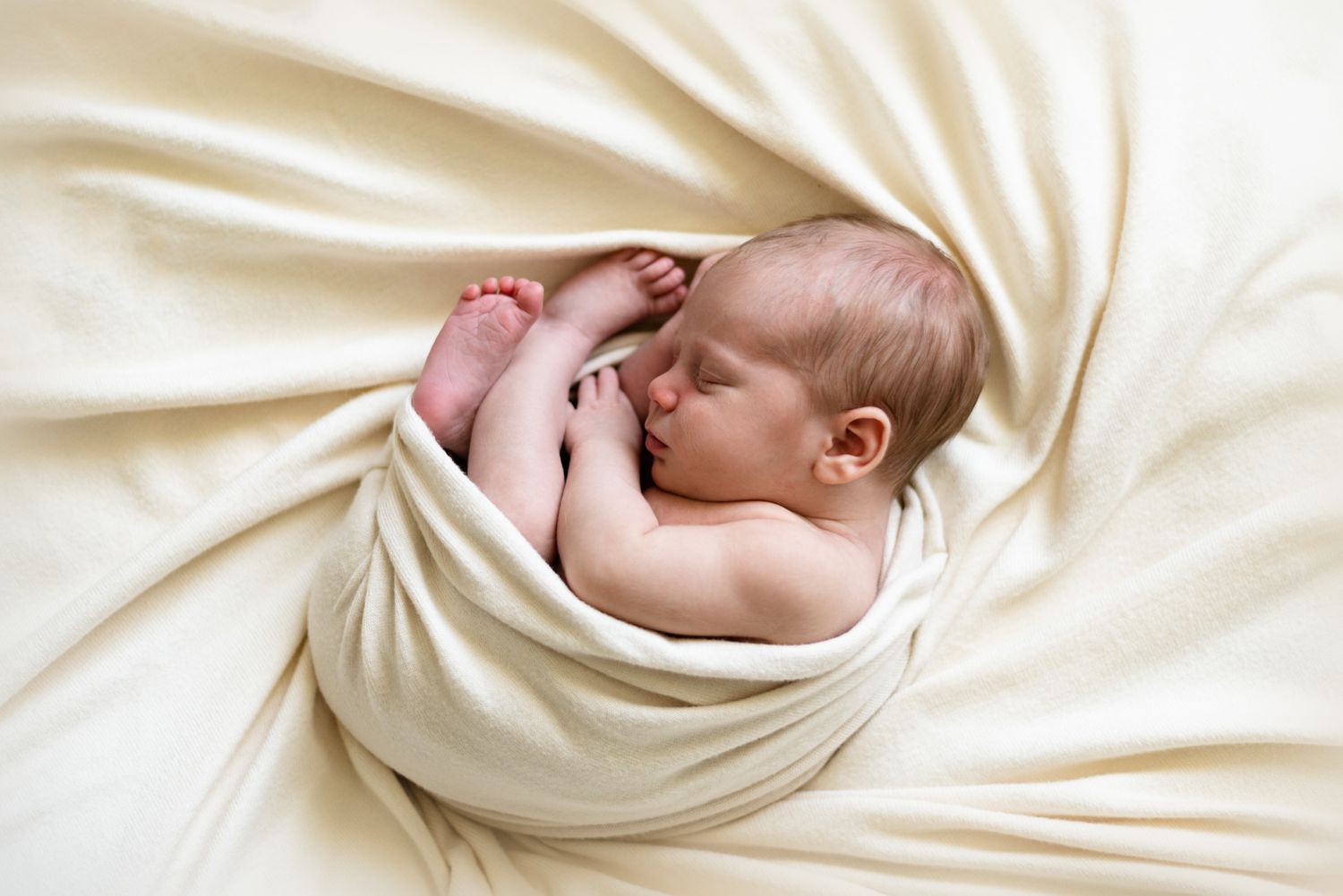 Sleeping baby nestled in cream colored wrap on light background.