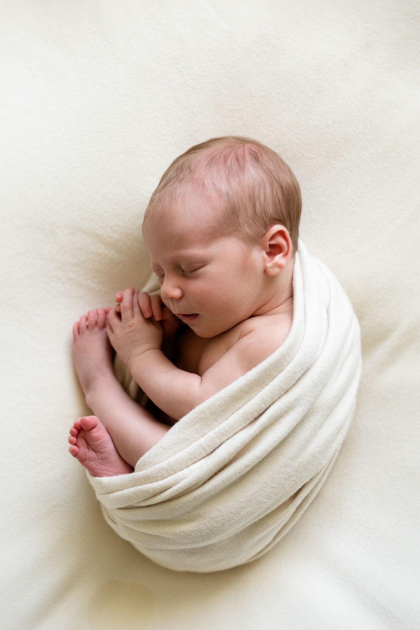 Peaceful newborn baby wrapped in soft white fabric sleeping in curled position.